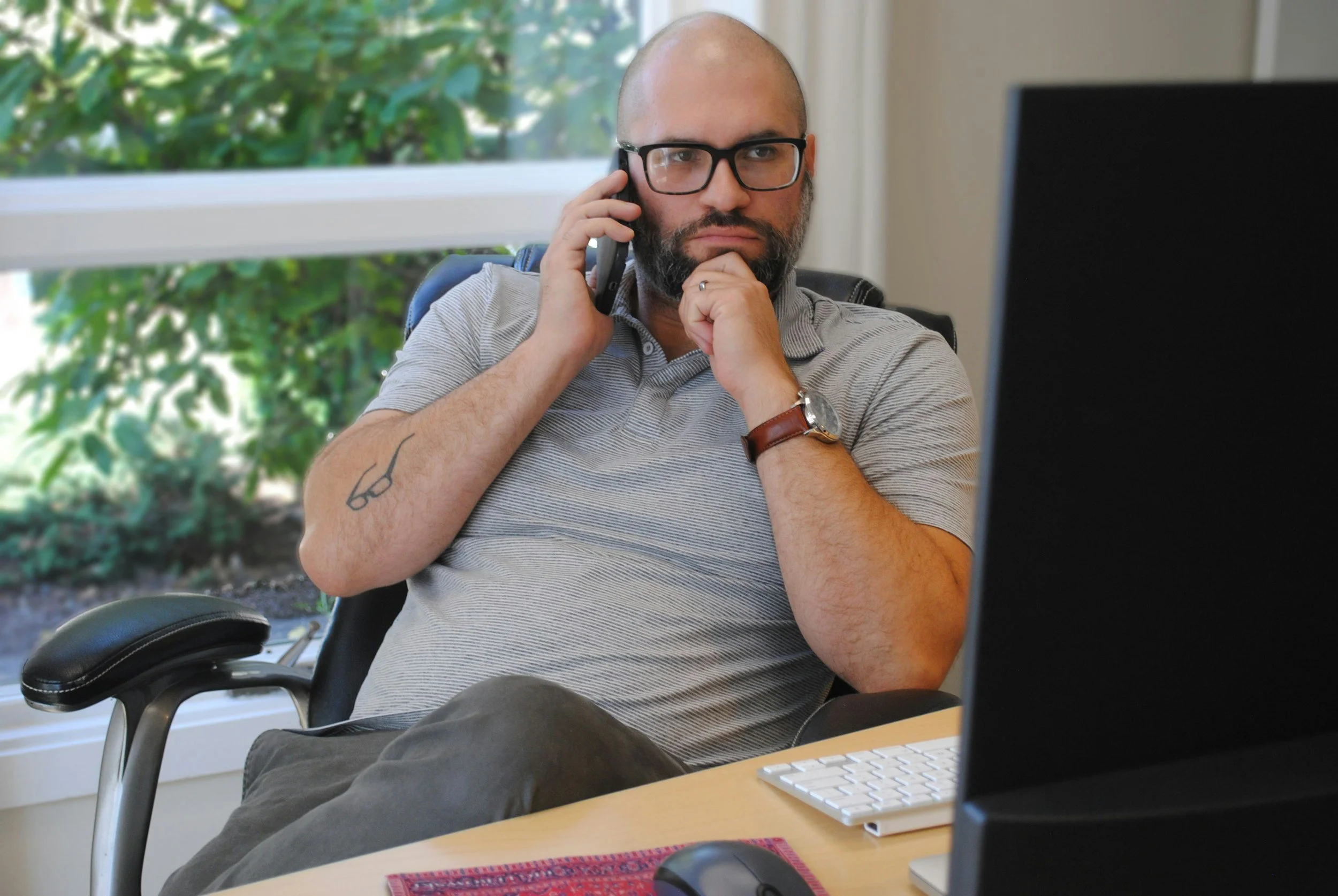 A bald man with glasses, a beard, and a tattoo of glasses on his arm is sitting at a desk, holding a phone to his ear with one hand and resting his chin on his hand, looking at a computer monitor.