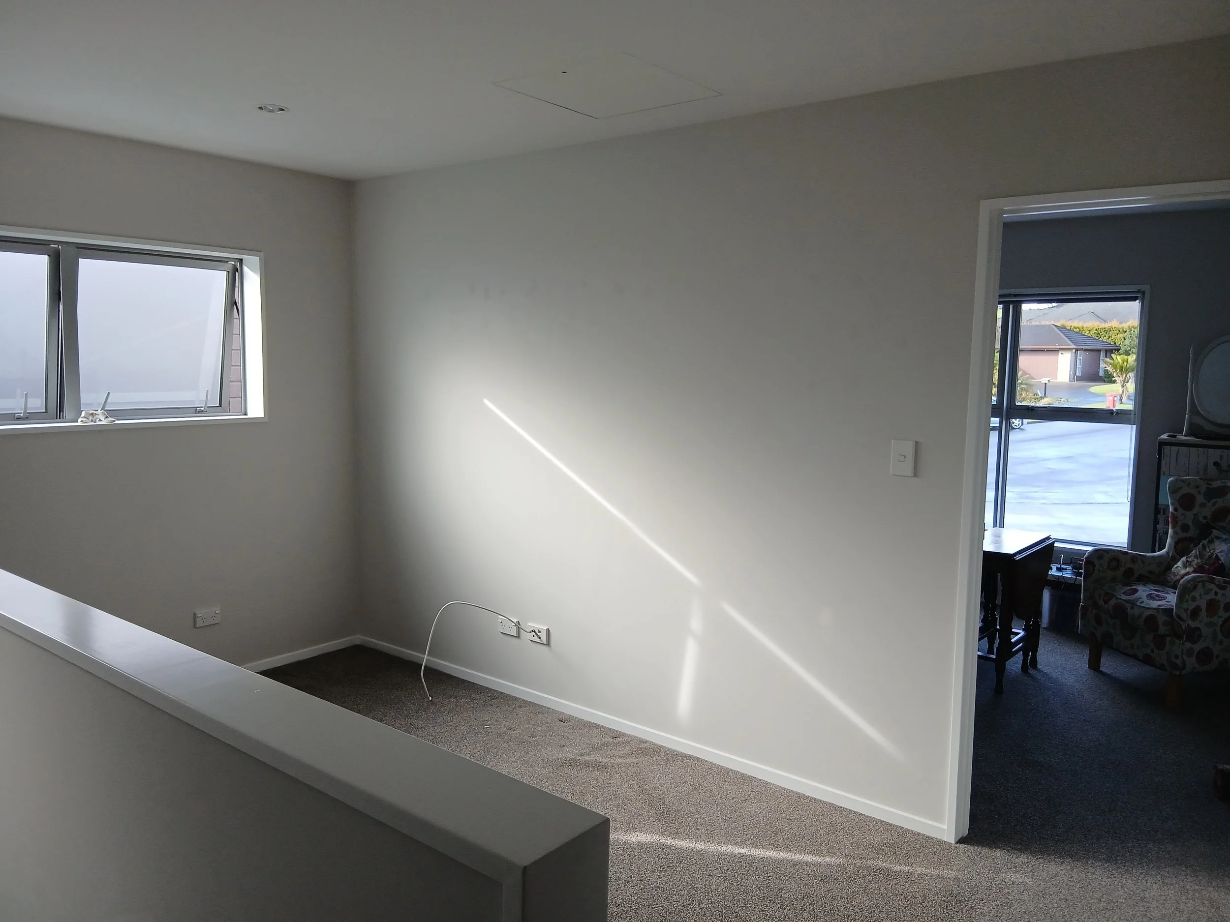 View of an empty, open plan room at the top of an internal stairwell with white walls, a window showing a house and street outside, and a doorway leading to a living area. Photo is taken in a two-storey home in Silverdale Auckland.