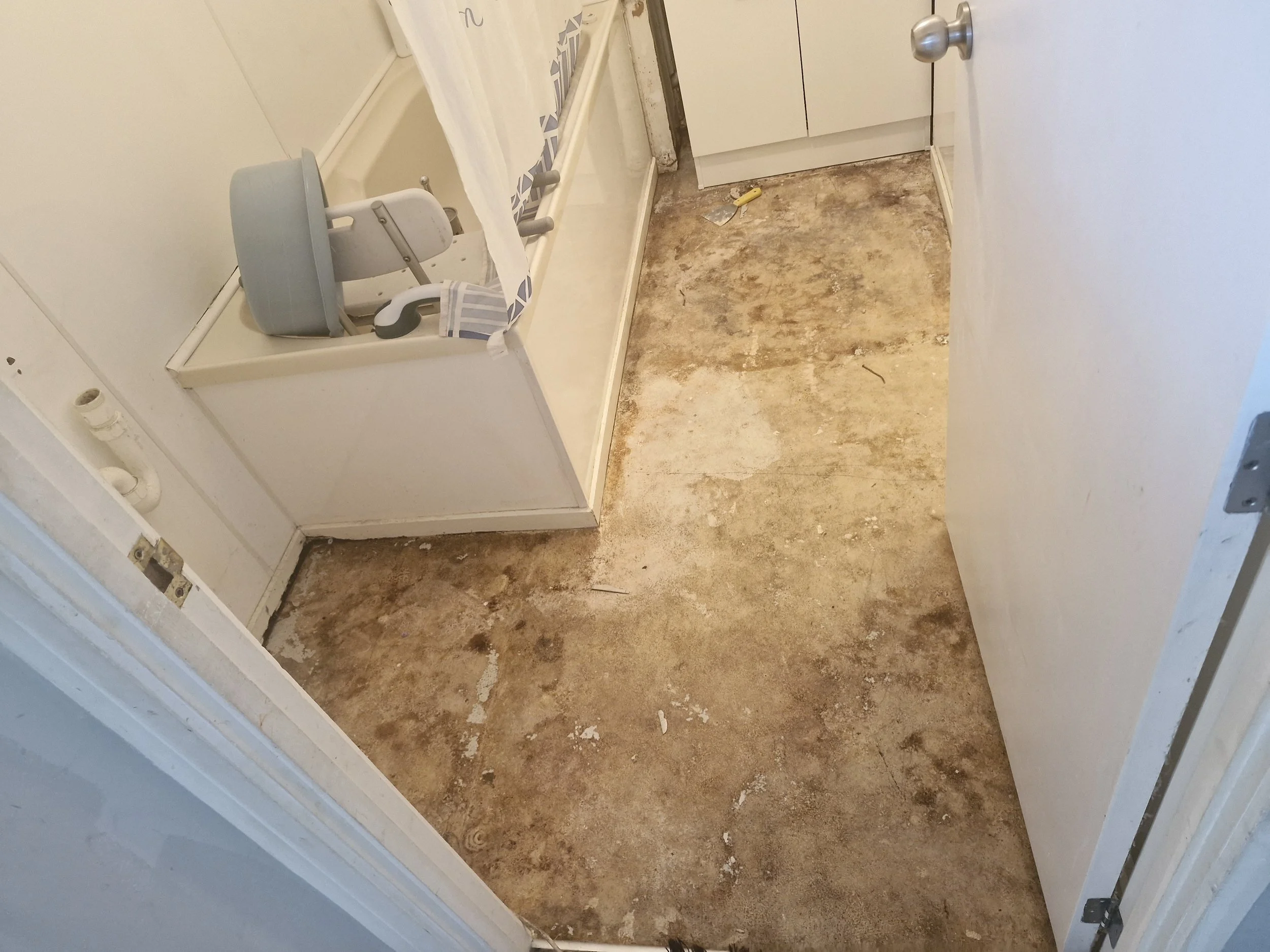 Bathroom with a partially removed floor showing subfloor, a white shower or bathtub with a shower curtain, a plastic child's high chair, a small white cabinet, and a door with a silver doorknob.
