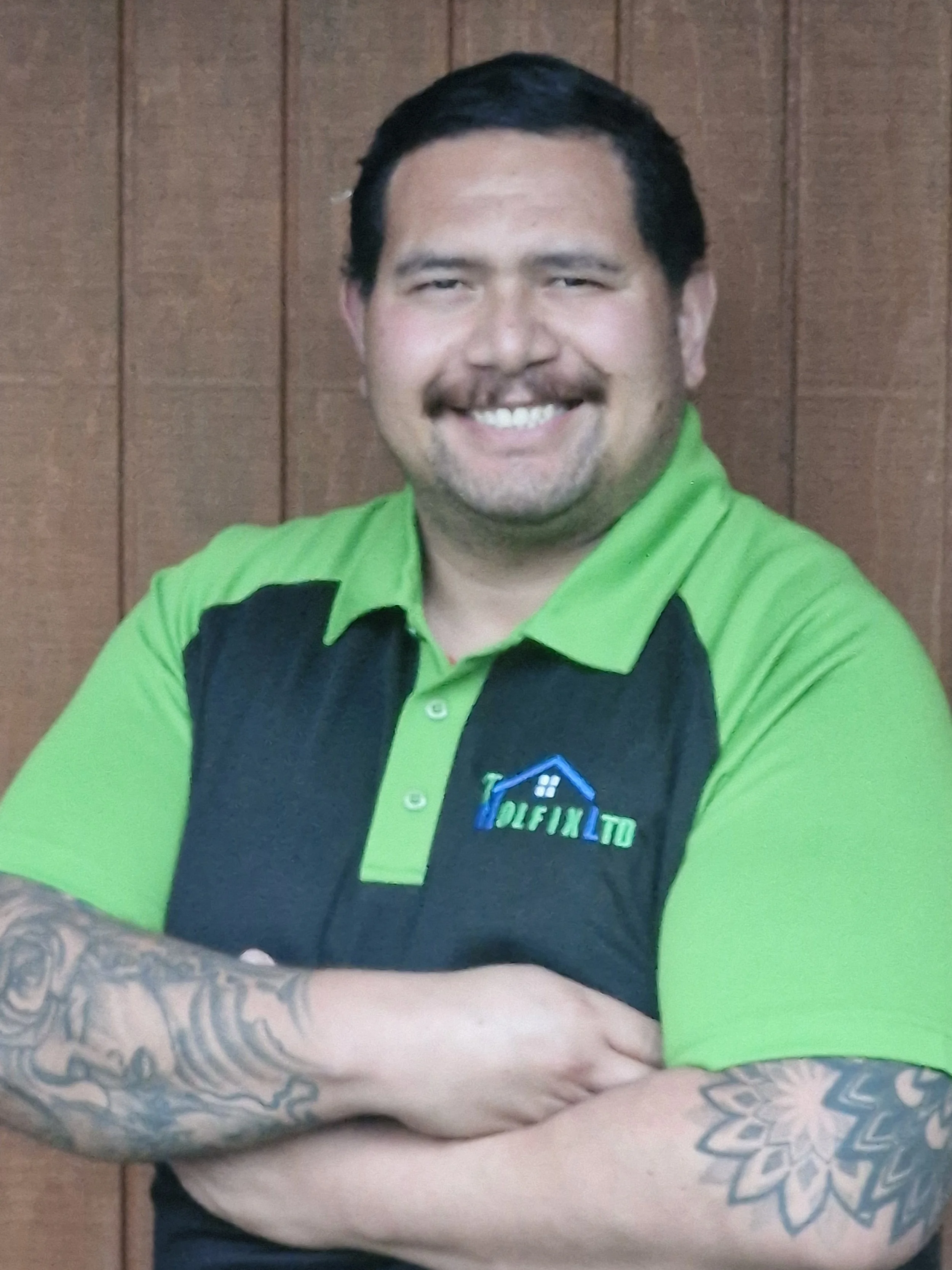Portrait image of Norton Shortland - a smiling man of māori descent with dark hair and moustache wearing a green and navy blue shirt,' has tattoos on his forearm, standing in front of wooden paneling.