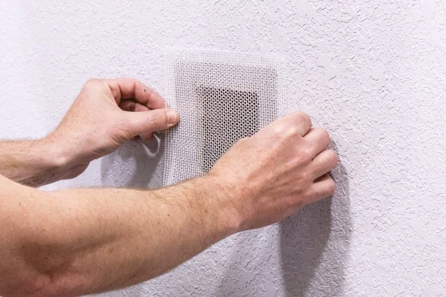 Hands installing a mesh cover over a hole on a white wall before commencing plaster repair work.