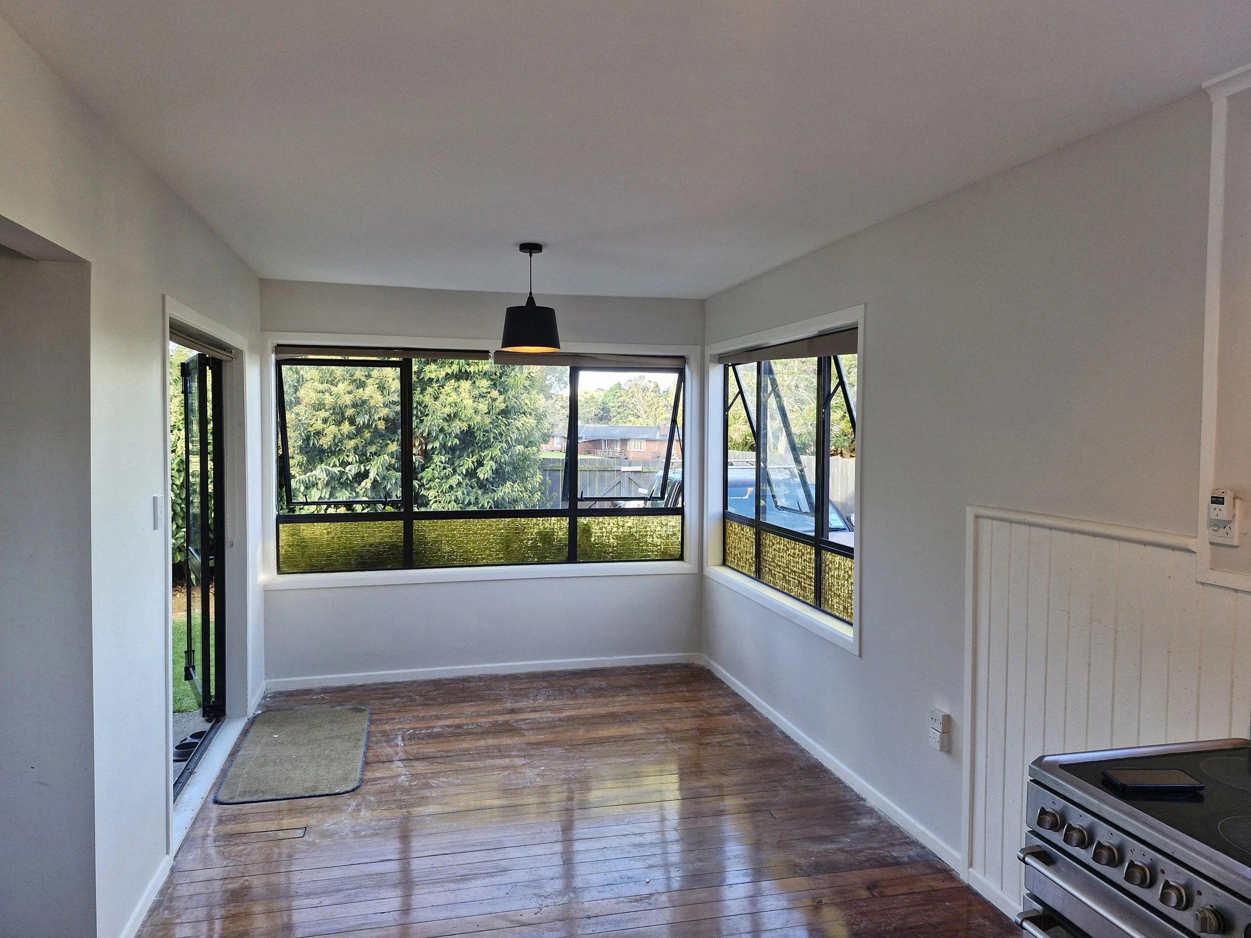 Empty dining area with hardwood floors, large windows, and a hanging black pendant light. Part of a kitchen with a stove visible in the corner.