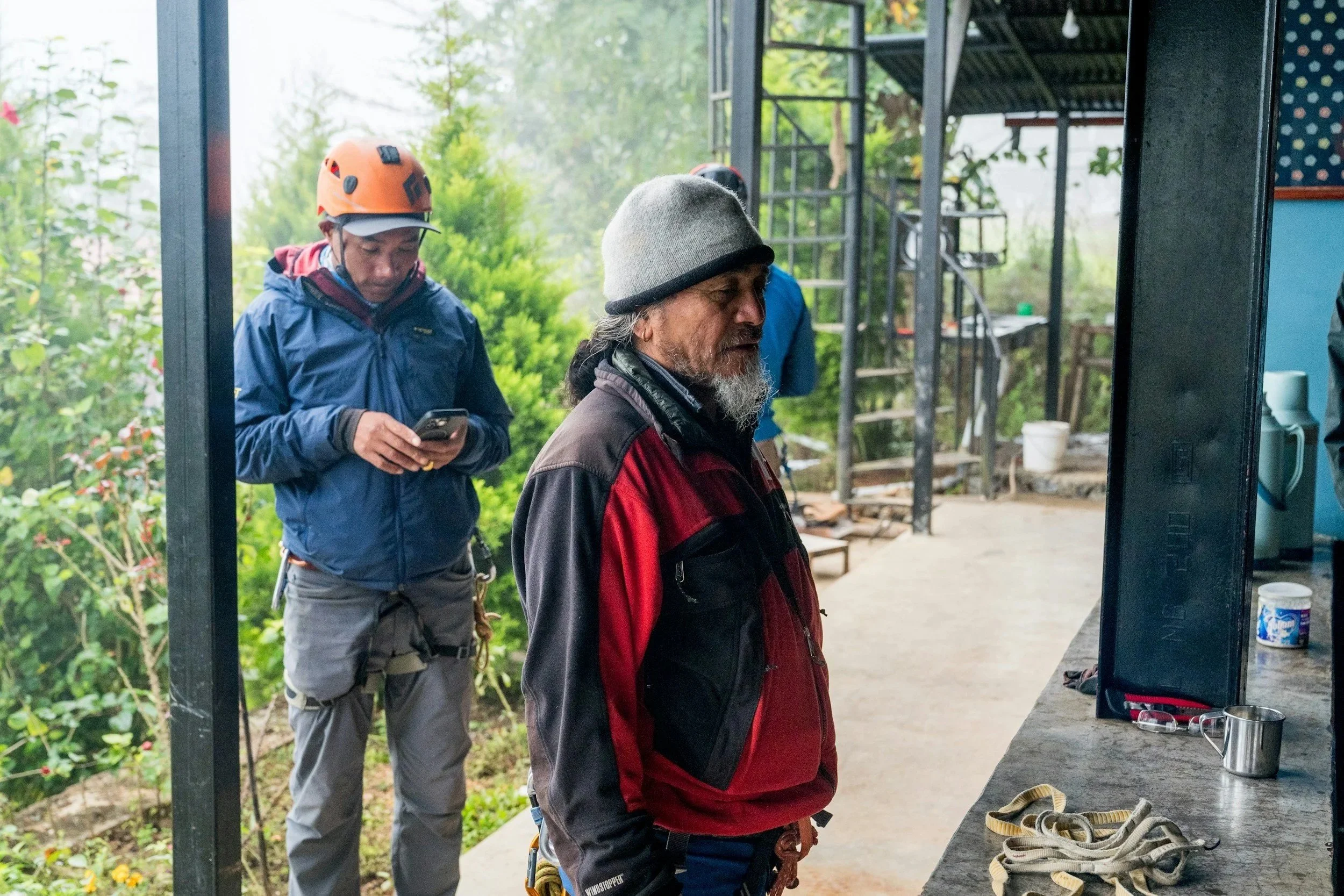 Two tradesmen standing outside.  One is wearing a red and black jacket with safety harness equipment. Second person in a blue jacket and wearing a hard hat looks at their phone. The setting is surrounded by greenery.