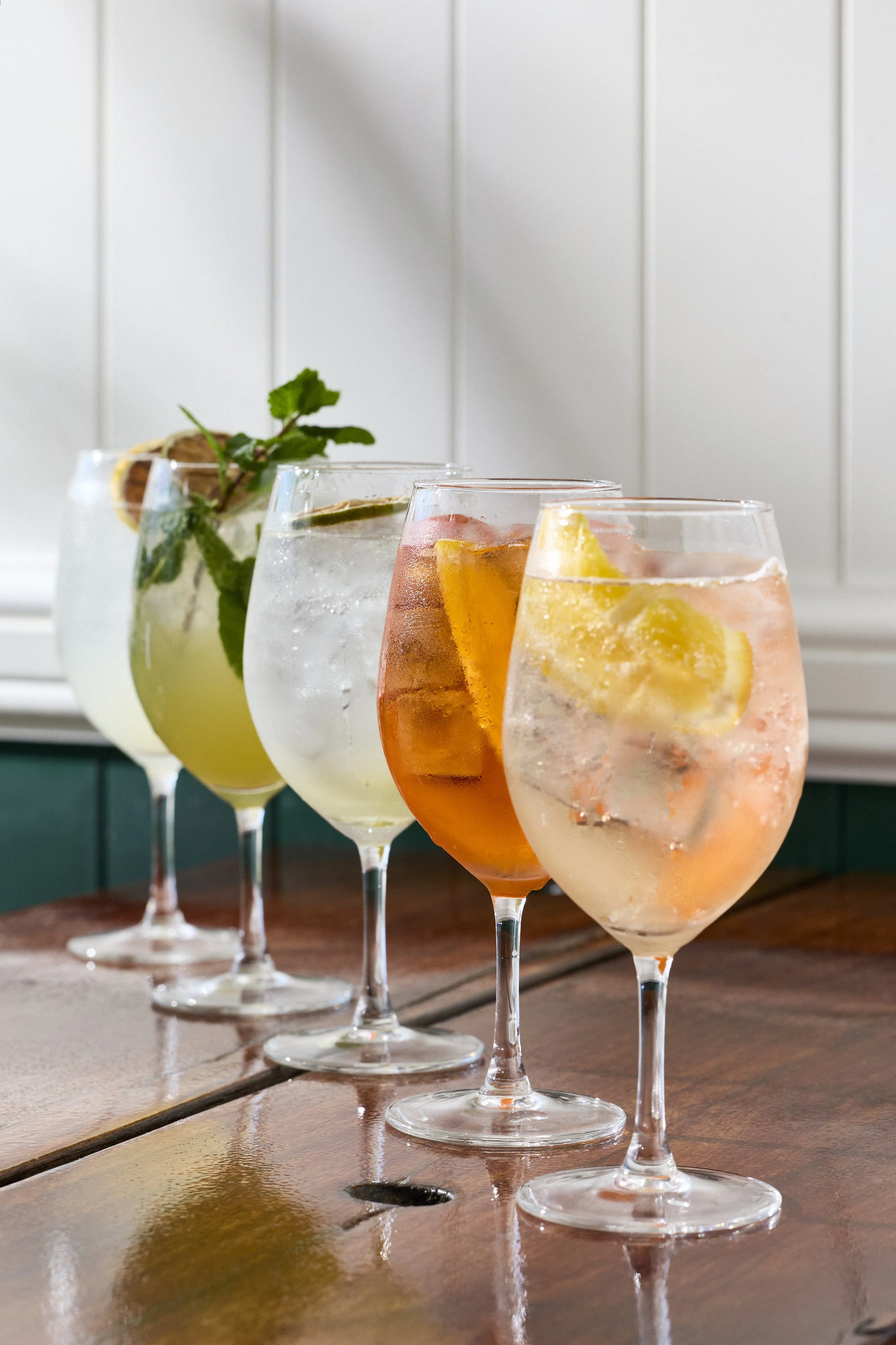 Four glasses of colorful cocktails with ice and fruit garnishes on a wooden table.