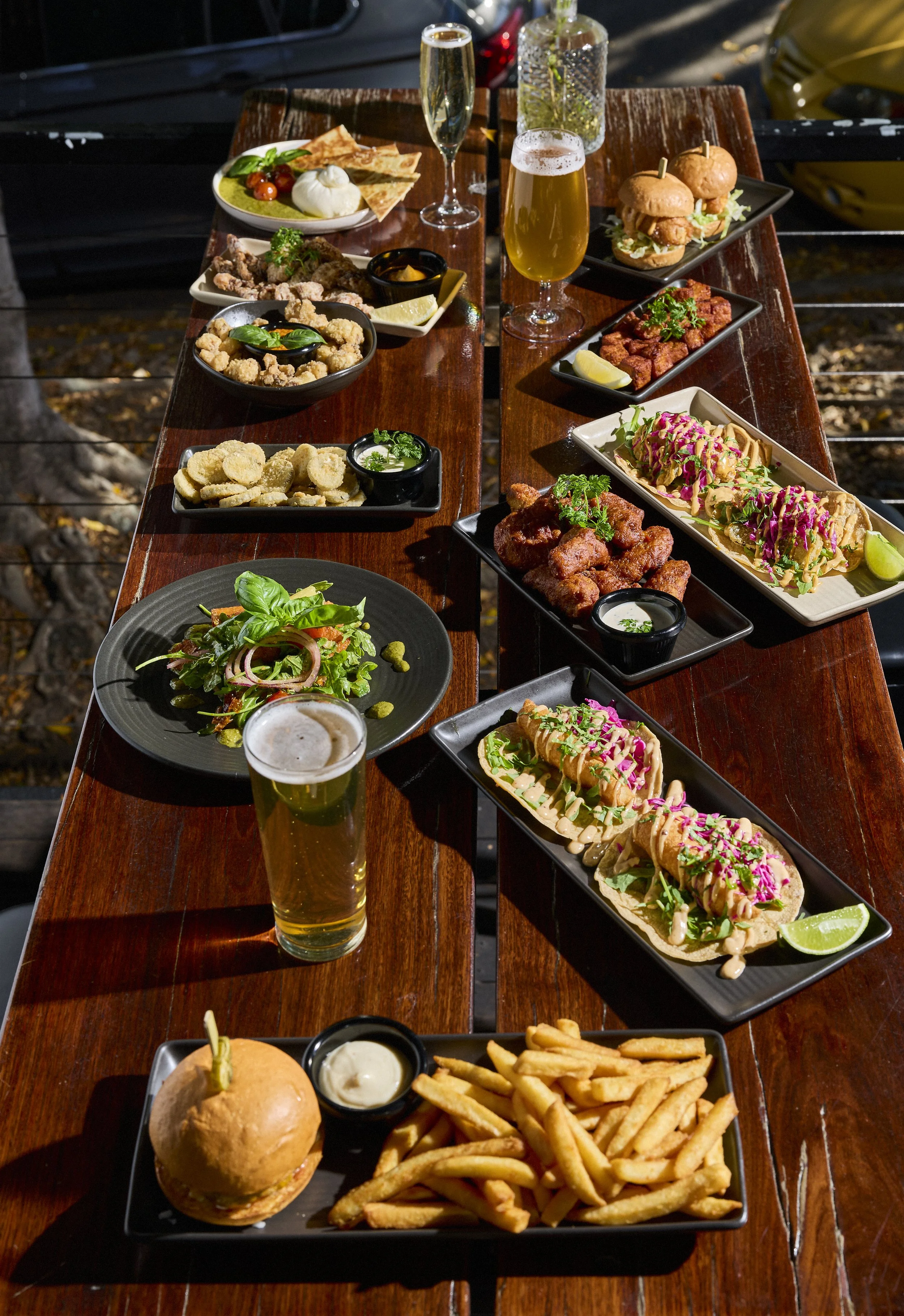 Outdoor dining table with various dishes including sliders, tacos, fried chicken, salad, fries, and drinks like beer, wine, and cocktails.