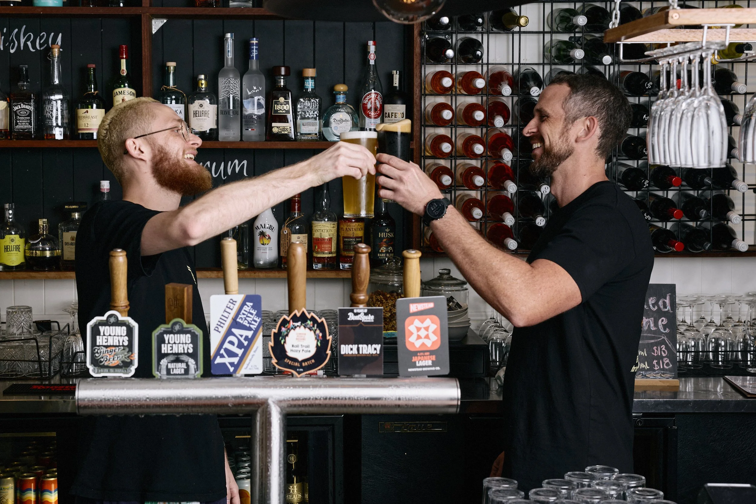 Two men at a bar toasting with glasses of beer, smiling. The bar has liquor bottles on shelves behind them and various beer taps on the counter.