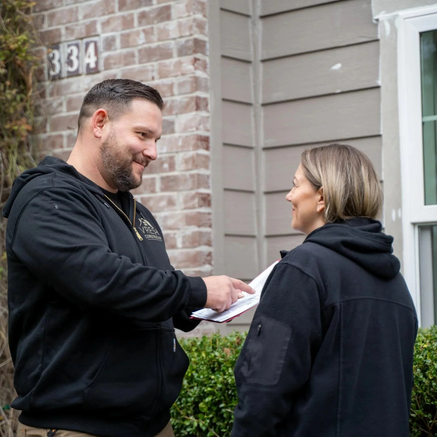 Contractor discussing plans with homeowner outside house, representing evaluation of egress window installation costs including foundation cutting, drainage, and window well requirements.