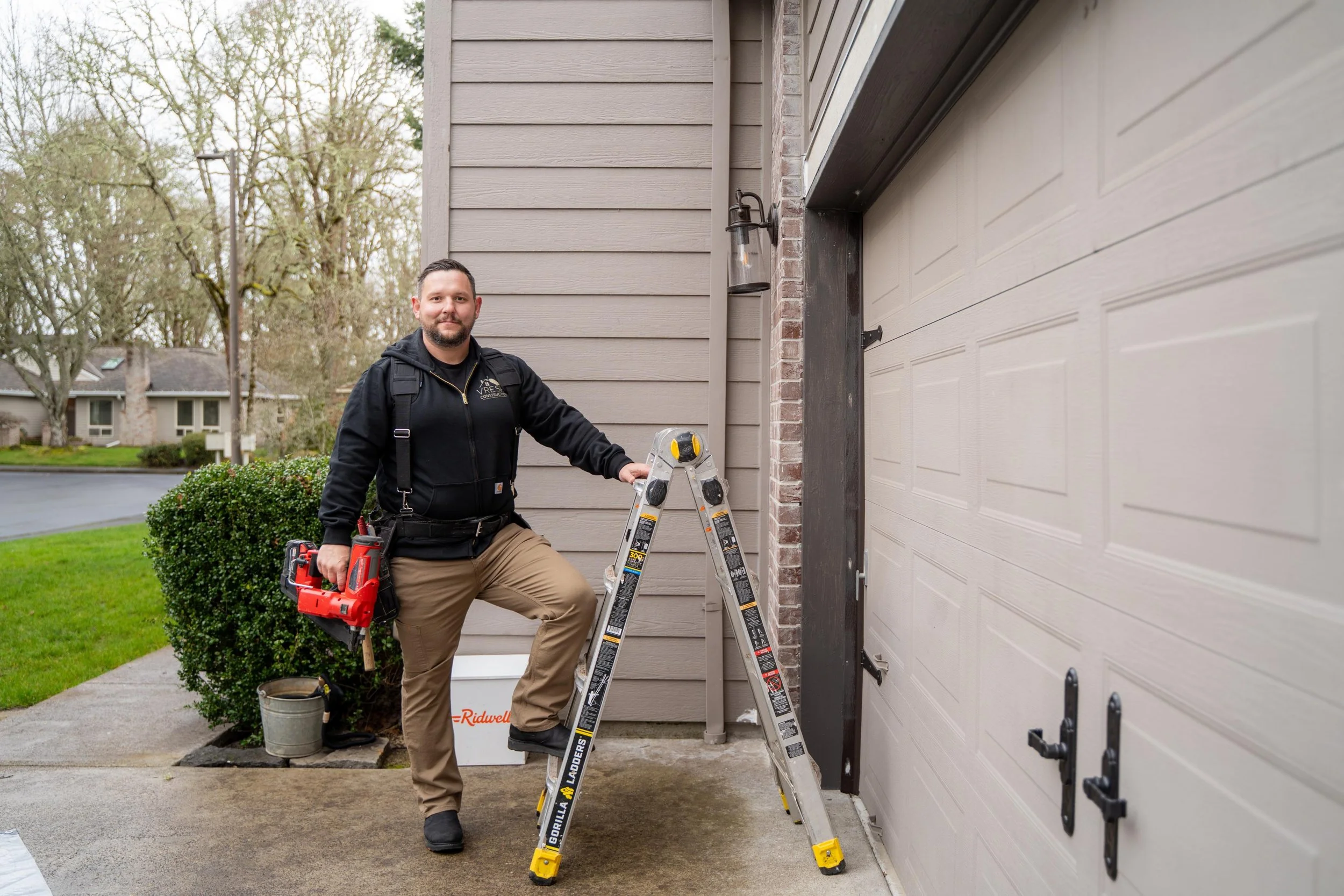 A handyman standing outdoors next to a ladder, holding a cordless power drill, in front of a garage door, with a house and trees in the background.