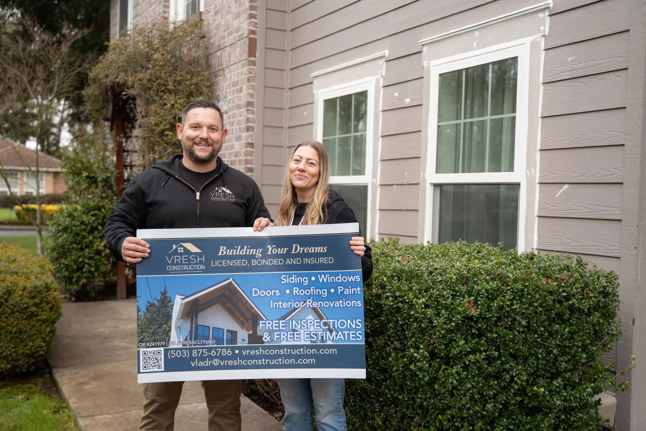 A man and woman standing outside near a house holding a sign for Vresh Construction, a home renovation company, smiling at the camera.