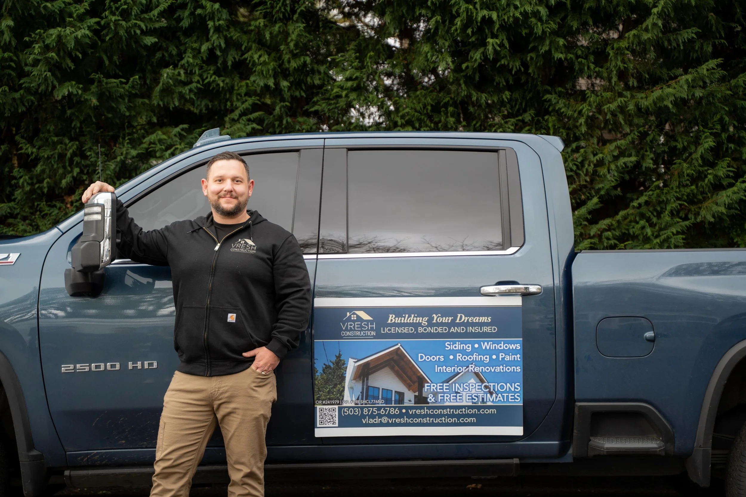 Man standing next to blue pickup truck with sign advertising VRESH Construction, holding an air impact wrench.