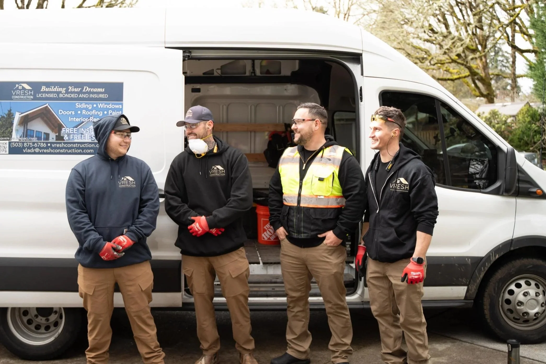 Four men in work clothes standing outside a white van, smiling and talking to each other. The van has a company logo and advertising on the side for construction and renovation services.
