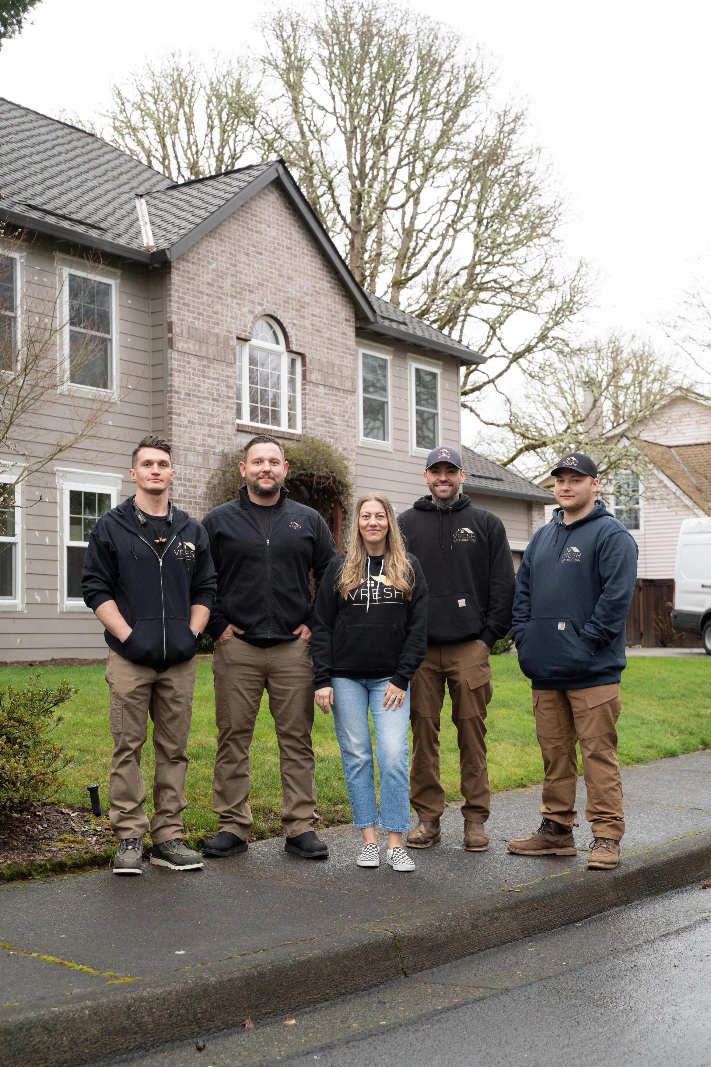 Five people standing on a sidewalk in front of a house, posing for a photo, with a lawn and trees behind them.