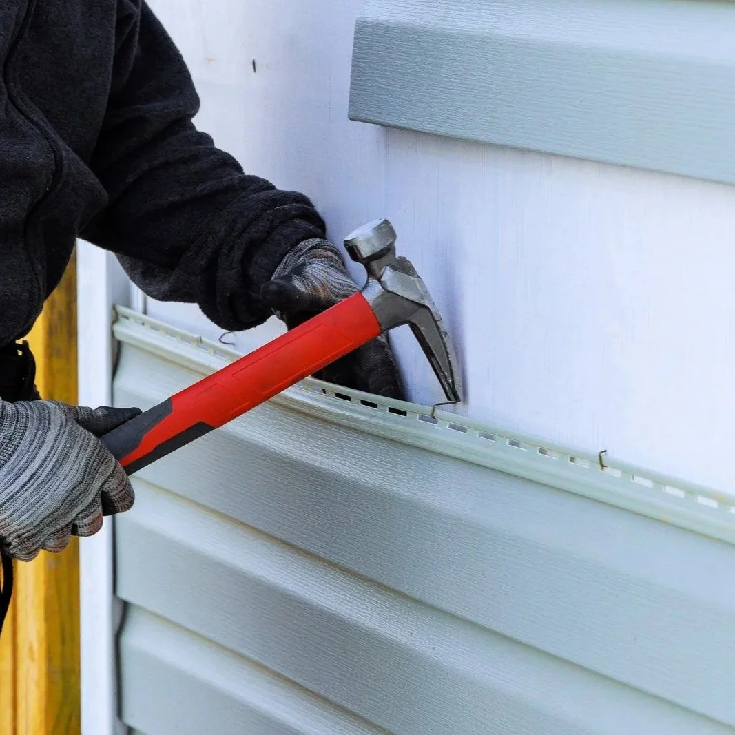 Close-up of gloved worker securing white vinyl siding panels using hammer on residential house exterior during installation work.