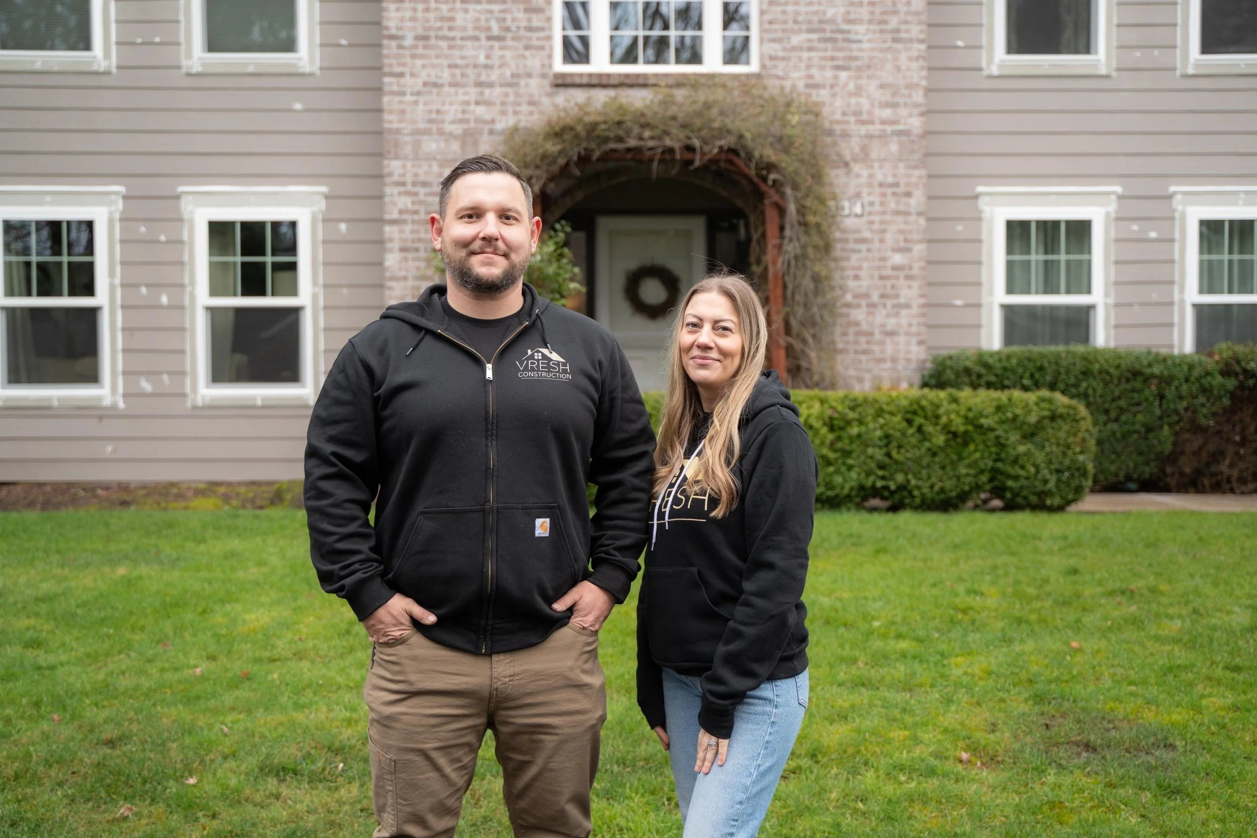A man and woman standing on a lawn in front of a house, both wearing black hoodies with a construction company's logo.