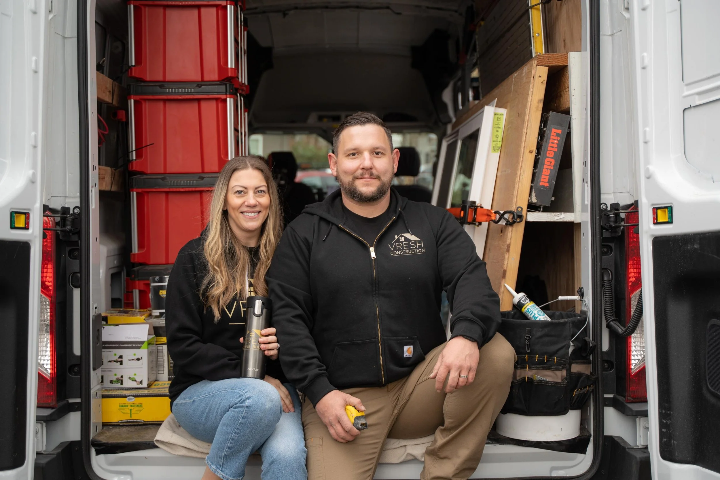 Two people sitting inside a packed delivery van, smiling at the camera. The woman on the left has long brown hair and is holding a black water bottle, while the man on the right has short dark hair and a beard, wearing a black hoodie with a construction company logo. The van's interior has red storage containers, a toolbox, and various tools and supplies.