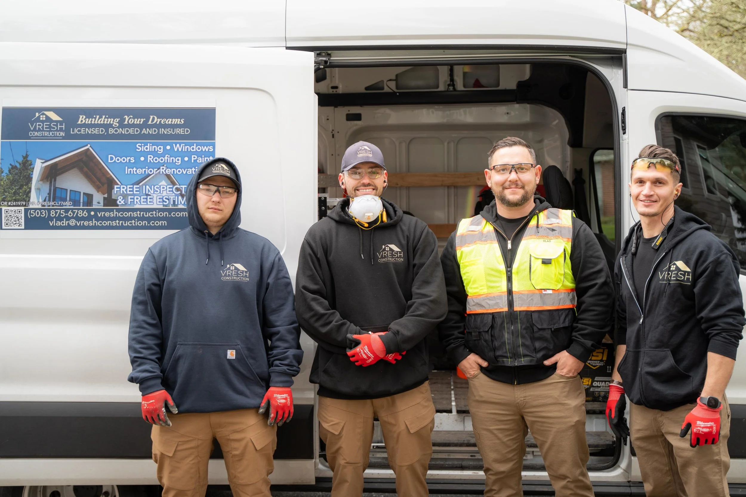 Four construction workers standing in front of a white work van with an open side door, wearing safety gear and branded clothing, smiling at the camera.