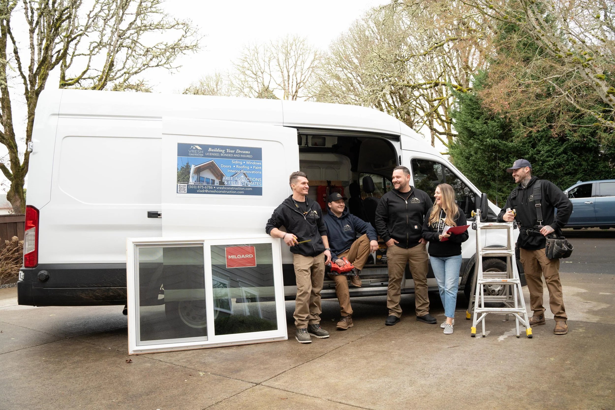 Group of five people standing and sitting in front of a white construction service van, with a ladder, window frame, and tools, outdoors on a cloudy day with leafless trees in the background.