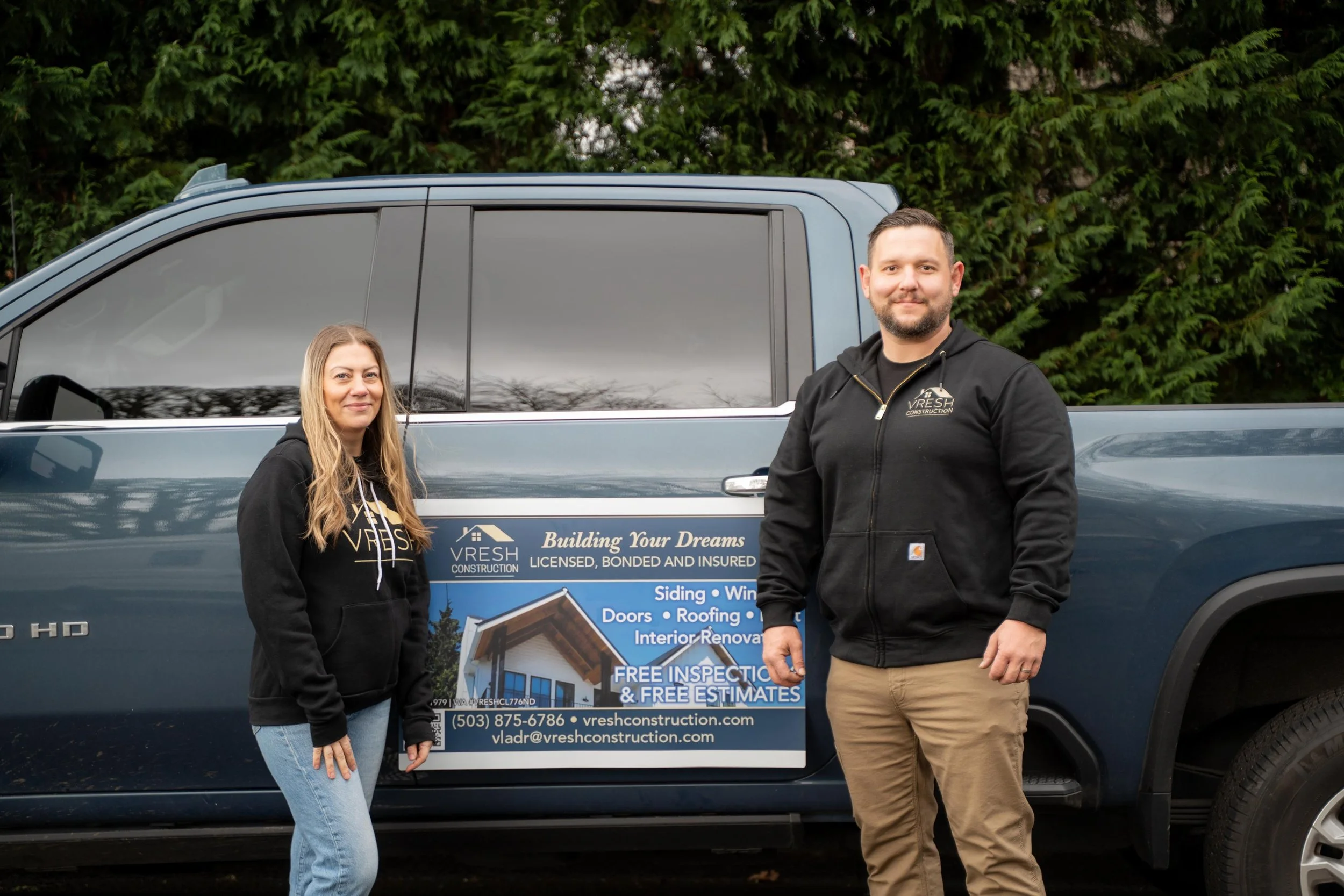 Two people standing next to a blue pickup truck with a promotional sign on its side, featuring the logo, contact information, and services of Vresh Construction company, with trees in the background.