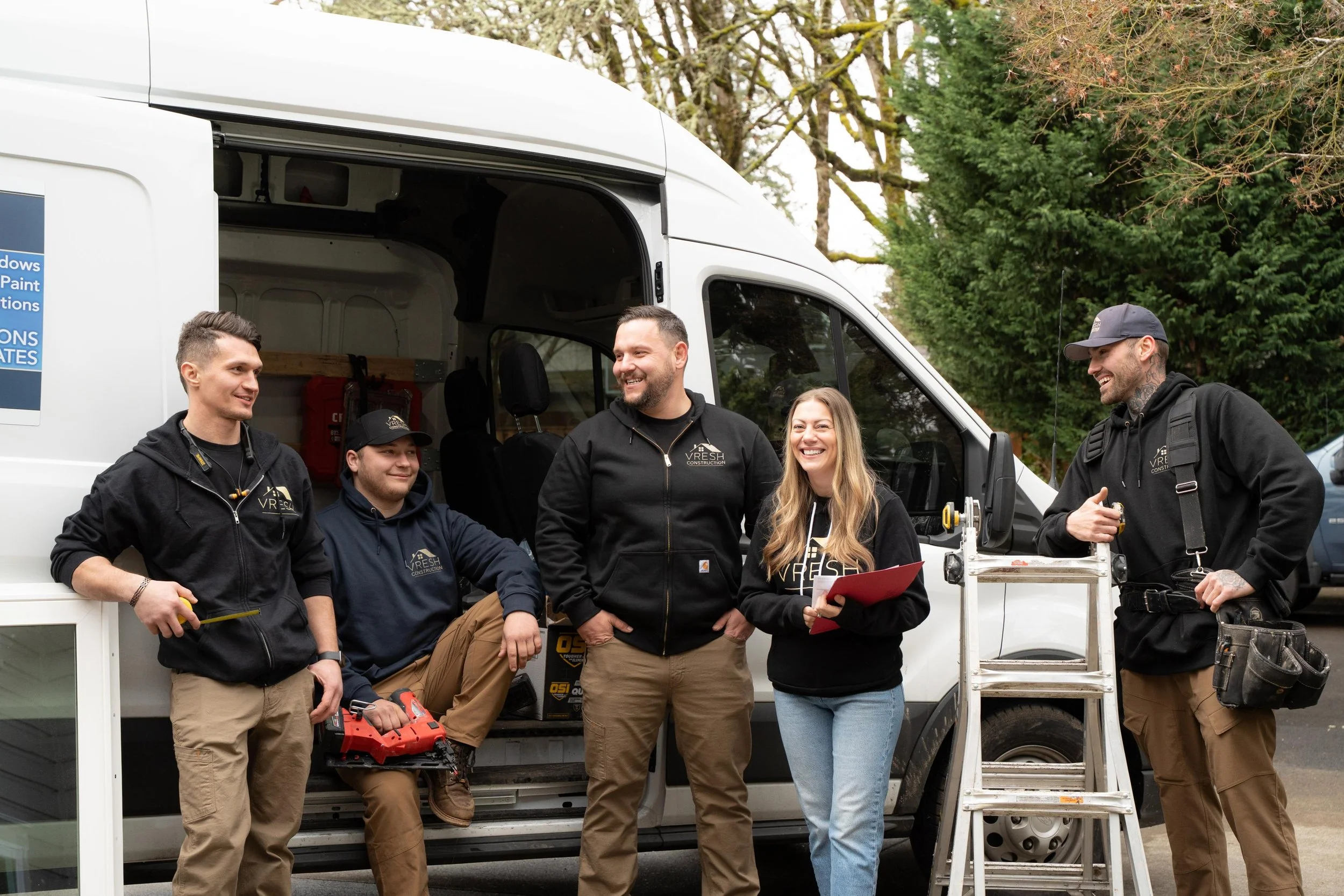Group of five construction workers and a woman talking and smiling standing near a white service van outdoors, with trees in the background.