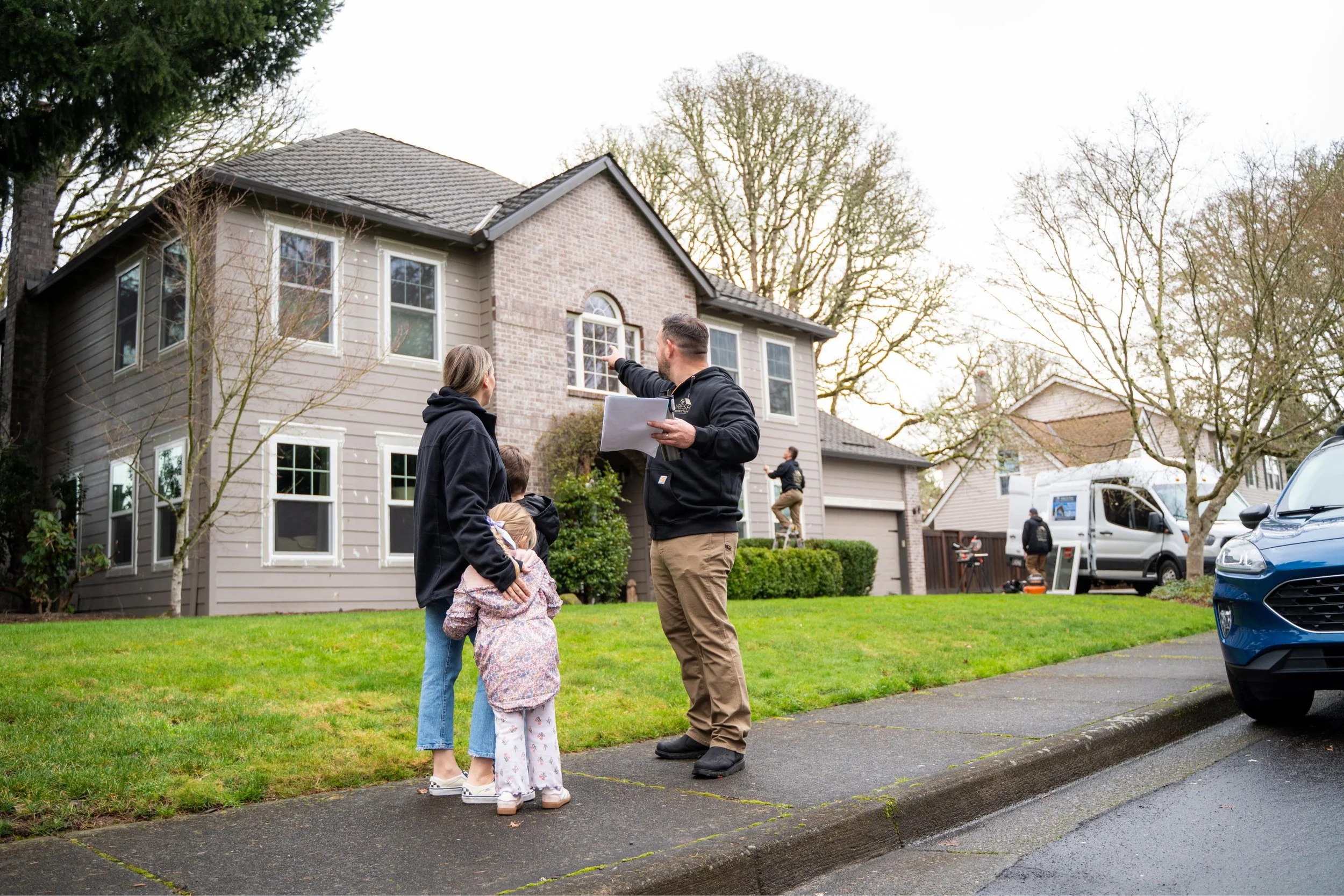 Real estate agent showing a house to a family on the neighborhood sidewalk. The agent is holding papers and pointing at the house, while the family listens. In the background, someone is working on the house's exterior. The scene is on a cloudy day with trees and parked cars.