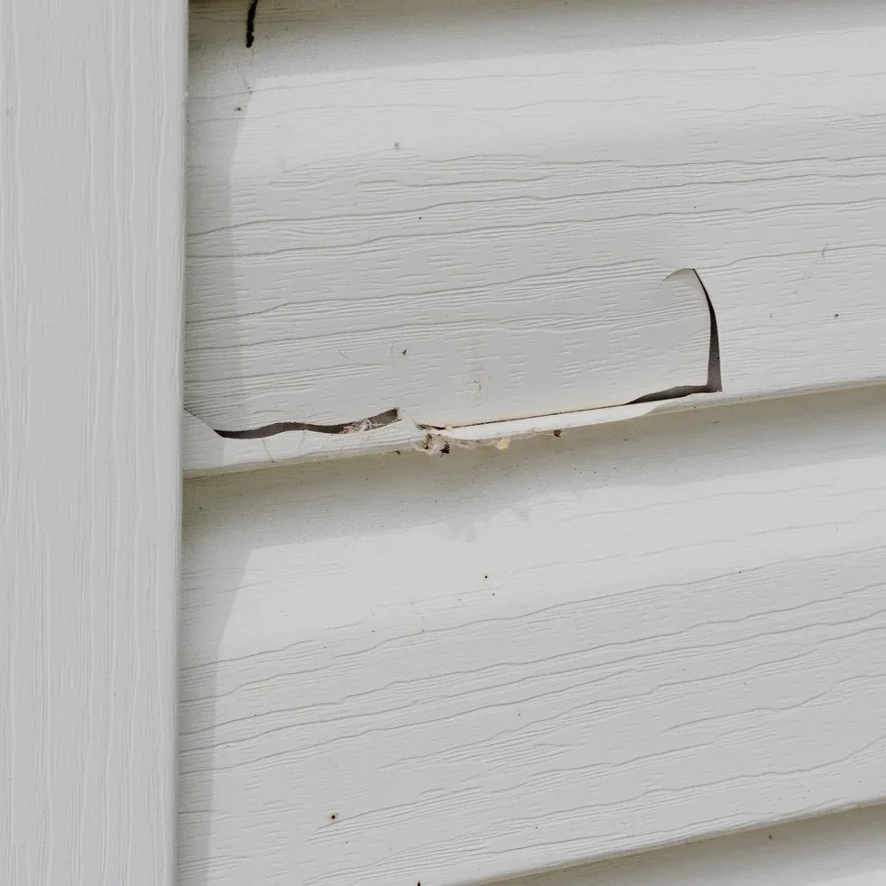 Close-up of cracked white vinyl siding panel with visible gaps allowing air infiltration, moisture intrusion, and exterior wall damage.