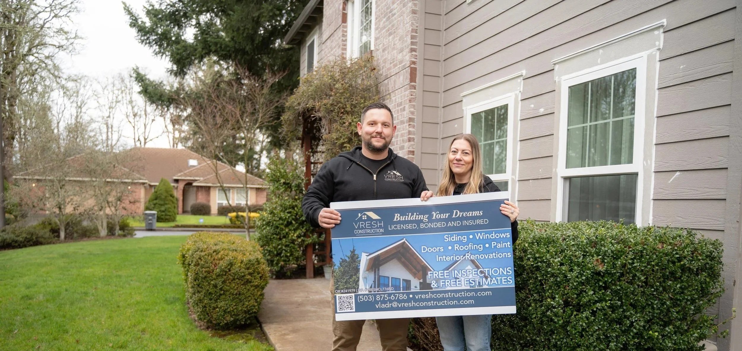 Two people standing outside next to a house, holding a sign advertising Vresh Construction services including siding, windows, doors, roofing, paint, and interior renovations, with a green lawn and neighboring houses in the background.