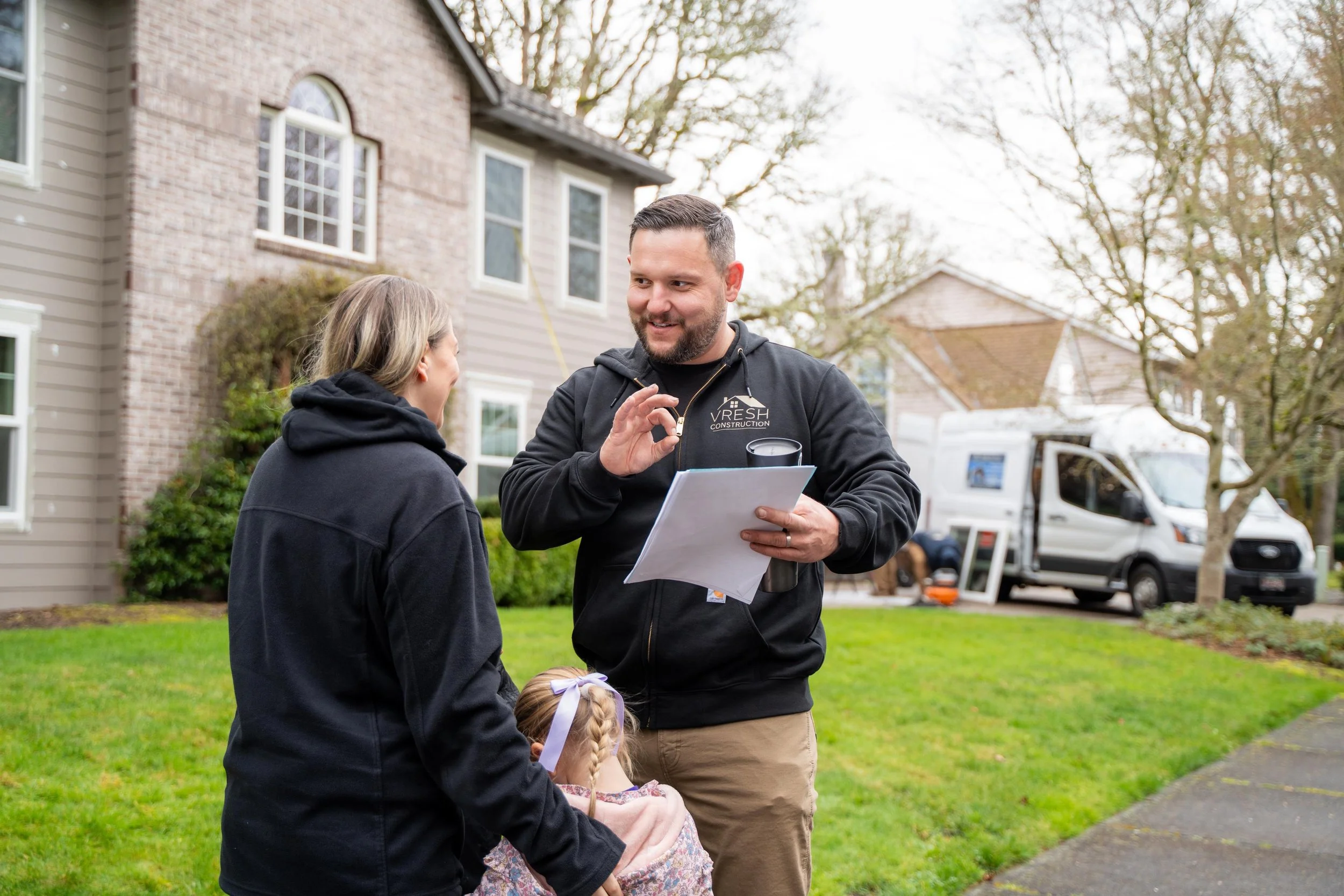 A construction worker talking to a woman and a girl outside a house with a yard and a moving truck in the background.