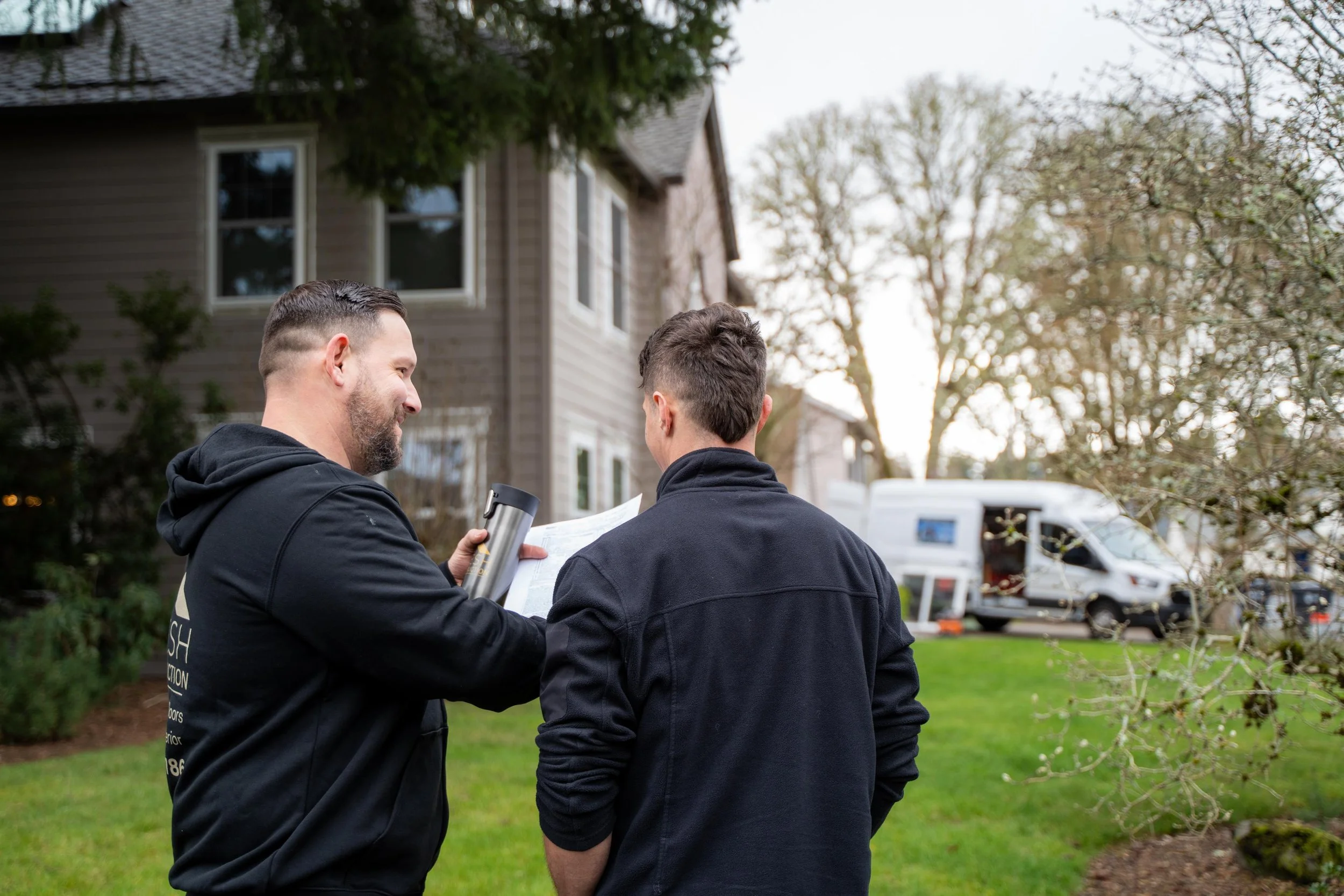 Two men having a conversation outdoors in front of a house, with a van parked nearby. One man is holding a water bottle and a clipboard.