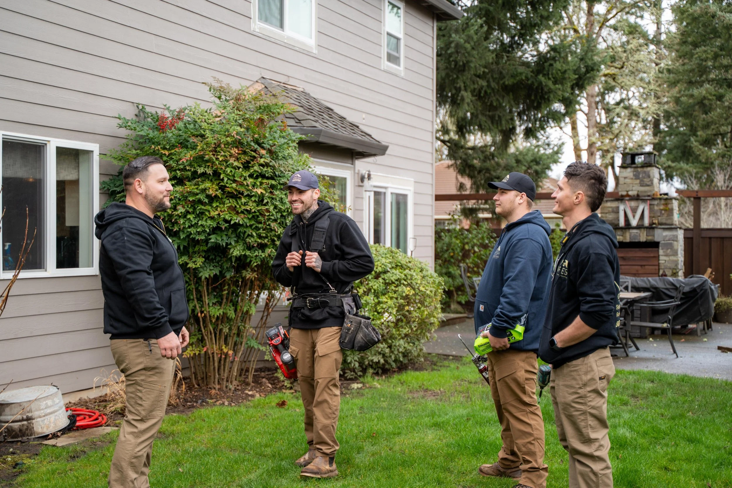 Four men in work attire standing outside near a house, engaged in conversation. They are holding tools, and there is a backyard with green grass, bushes, and outdoor furniture in the background.