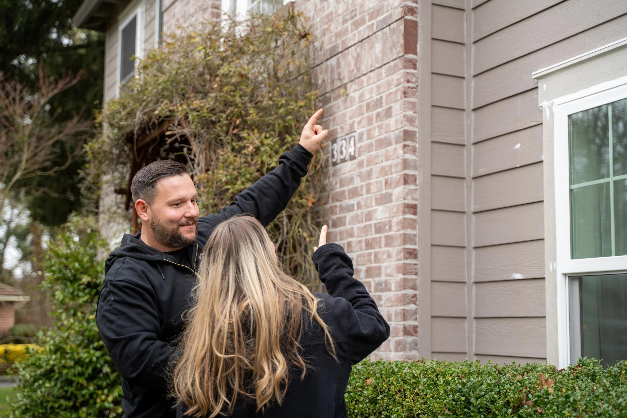A man and a woman pointing to a house number on a brick wall outside a house.