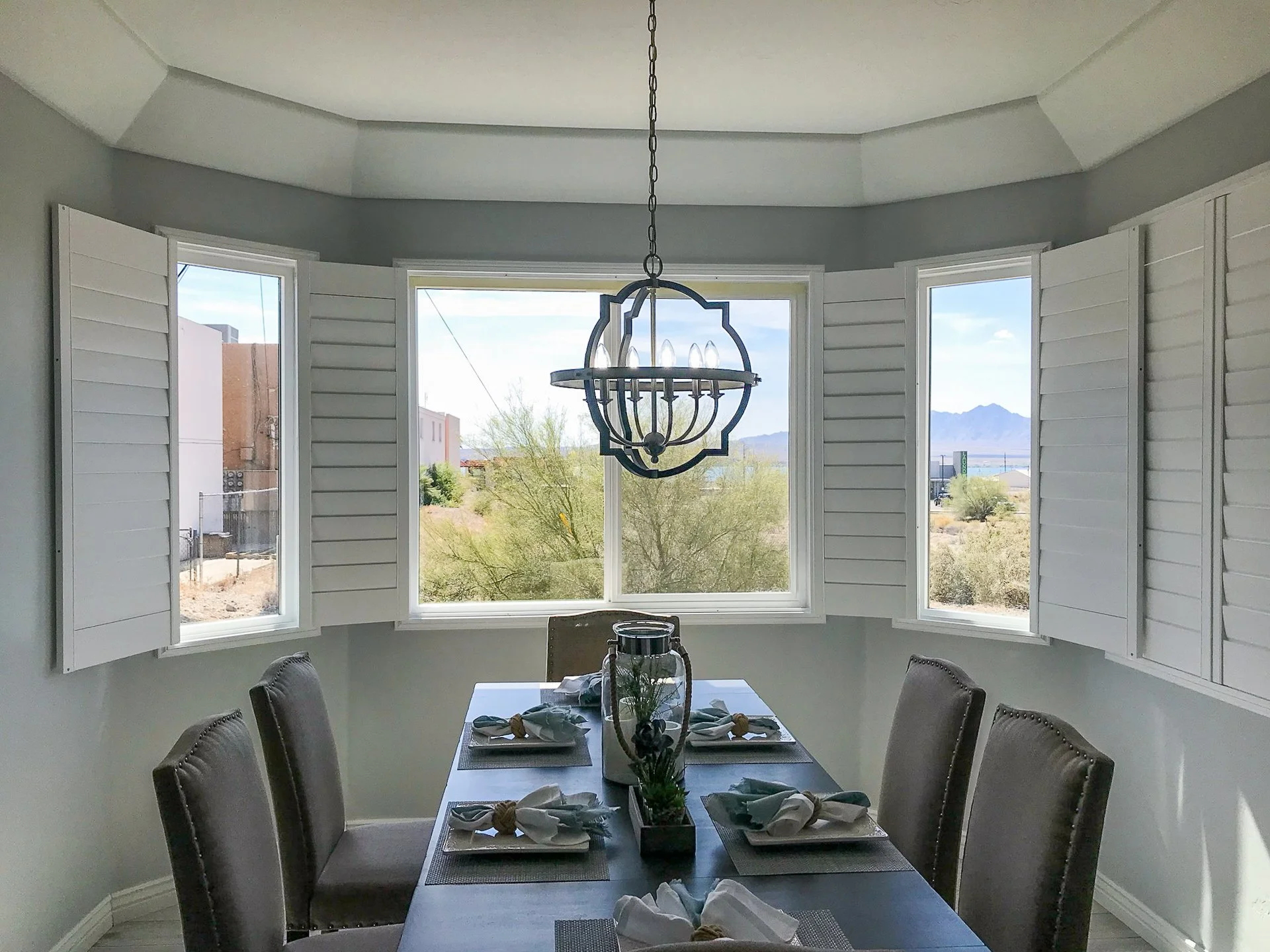 Dining room with a dark wooden table, six upholstered chairs, a centerpiece, and a modern chandelier, with large windows showing a desert landscape and mountains in the distance.
