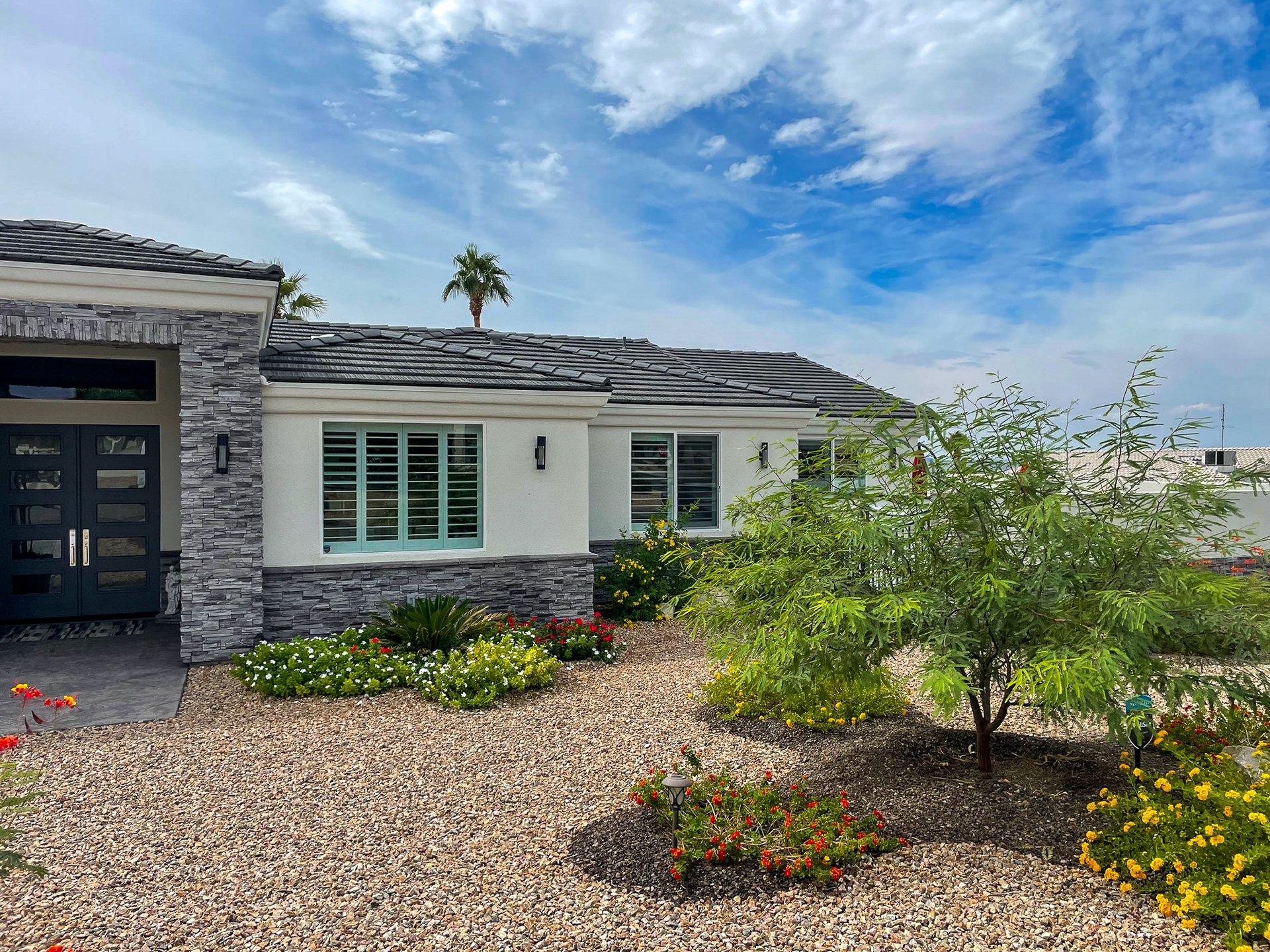 Front view of a modern house with white walls, gray stone accents, a black front door, and a garden with various colorful flowers and plants under a partly cloudy blue sky.