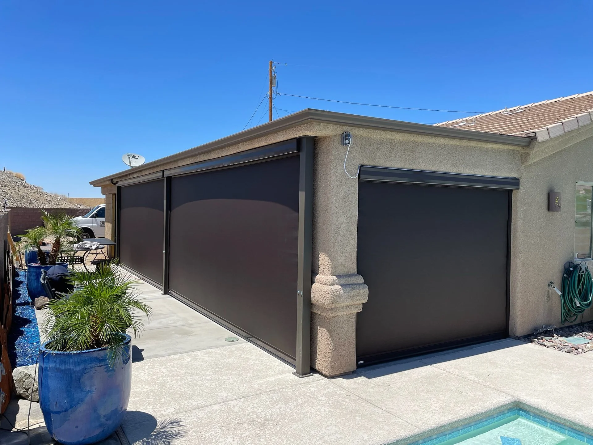 A house with a screened-in porch and a closed black garage door. There are potted plants and a small swimming pool in the backyard, along with a hose reel on the wall.
