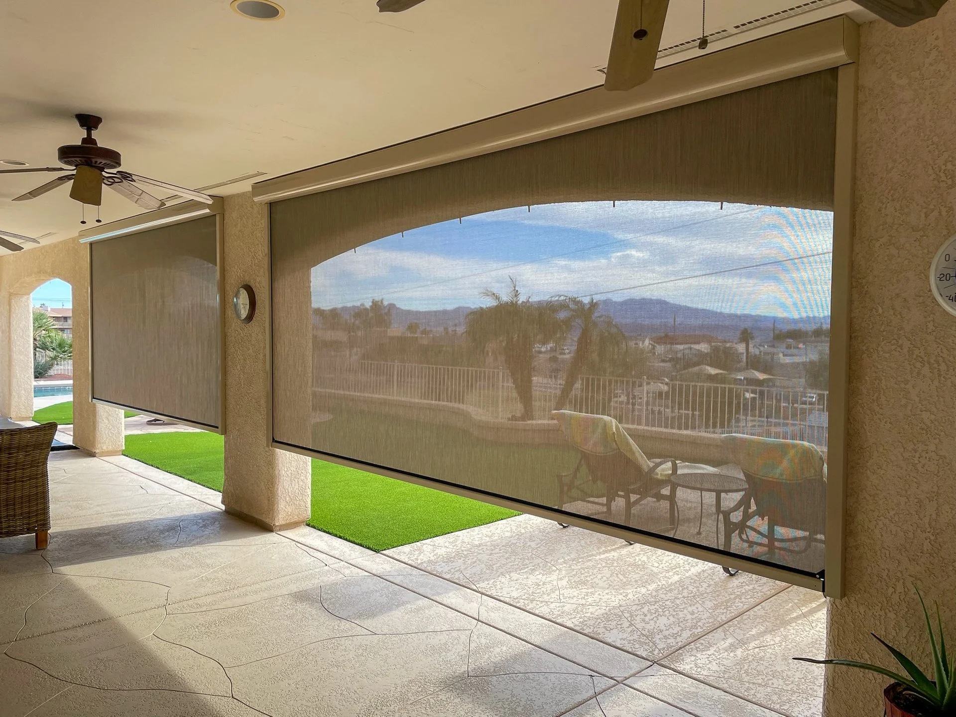 Back patio with two large screened sliding doors, ceiling fans, outdoor chairs, and a view of a backyard with green grass, trees, and distant hills under a partly cloudy sky.
