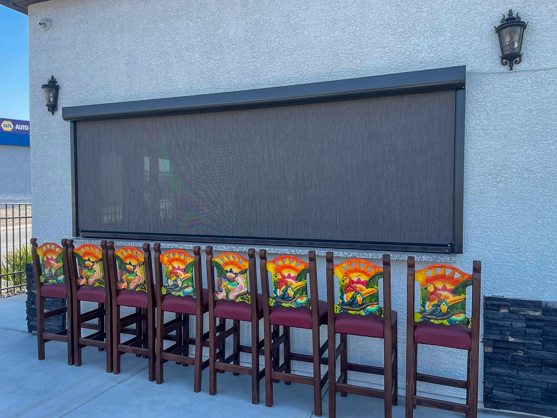 Seven colorful, decorated chairs with a sunset and bird design sit in front of a blank outdoor movie screen mounted on a white wall, with two outdoor lanterns on each side.