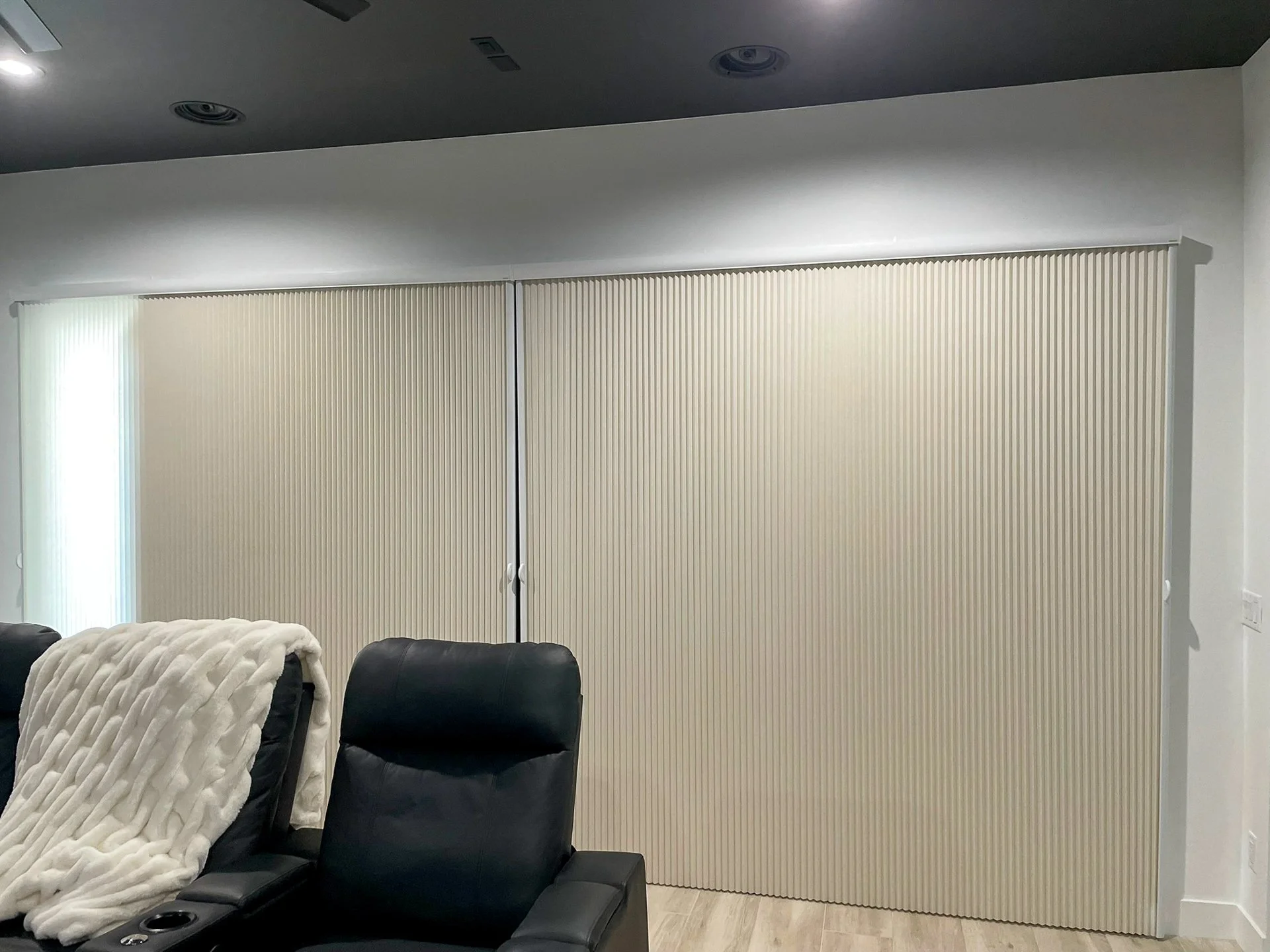 A living room with black leather chairs, one with a white fleece blanket, and closed beige vertical blinds covering large sliding doors or windows, with a wooden floor and a white wall.
