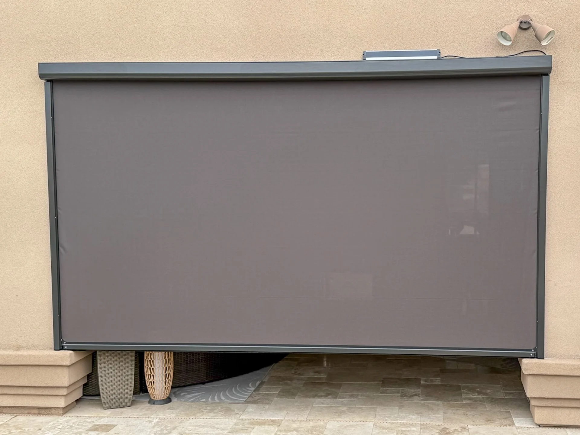 A gray retractable outdoor privacy screen mounted on a beige wall, with outdoor lights above and patio furniture underneath.