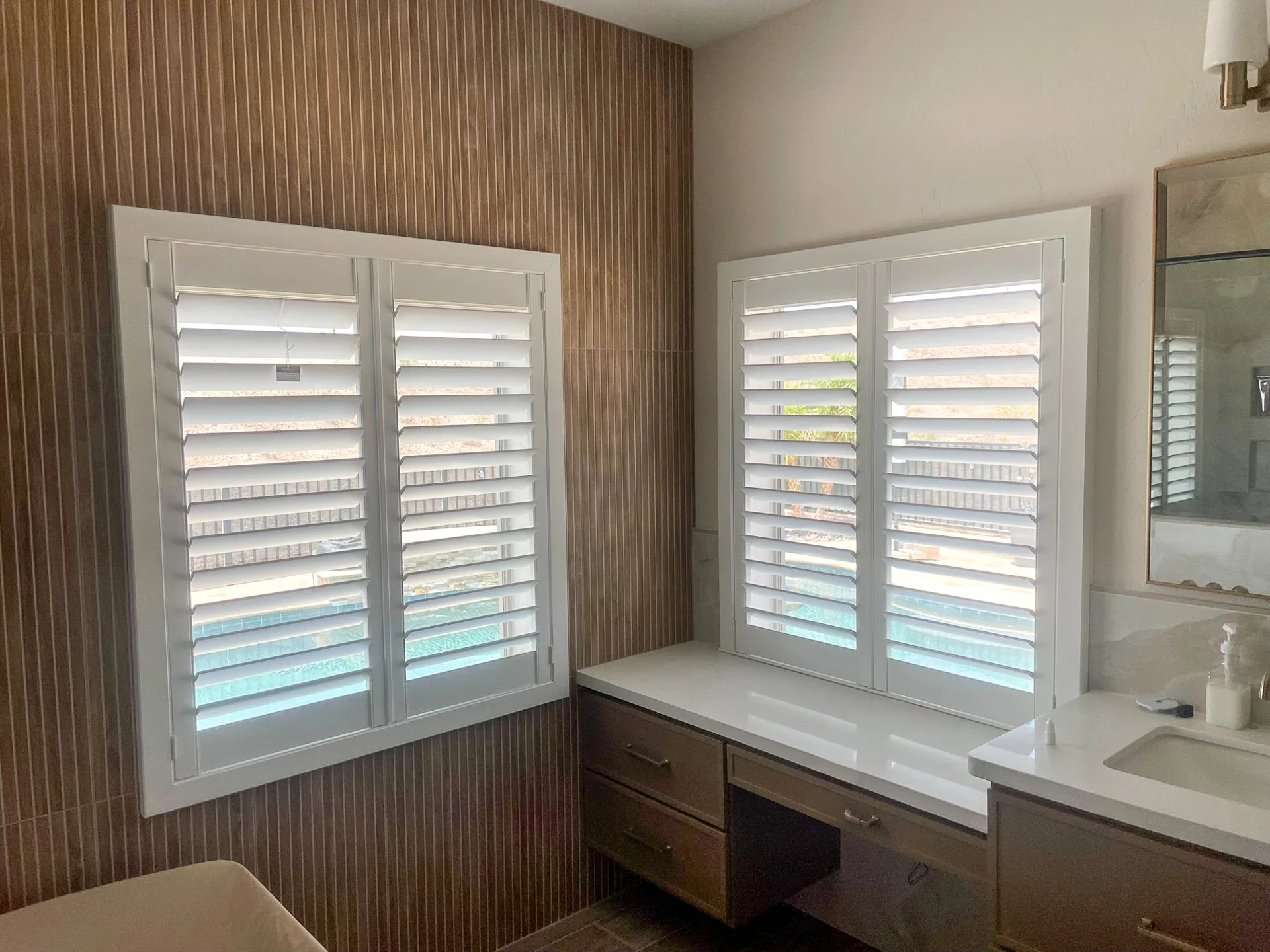 Bathroom with wooden wall and two windows with white plantation shutters, a white countertop, and a mirror.