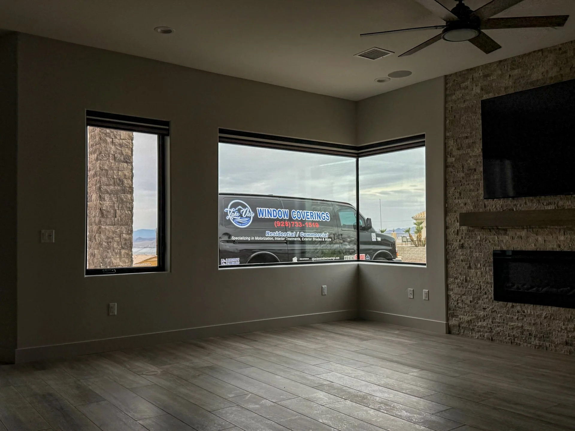 Empty living room with large windows showing a vanAdvertisement for window coverings outside, white walls, wood flooring, a ceiling fan, a stone fireplace area, and a mounted TV.