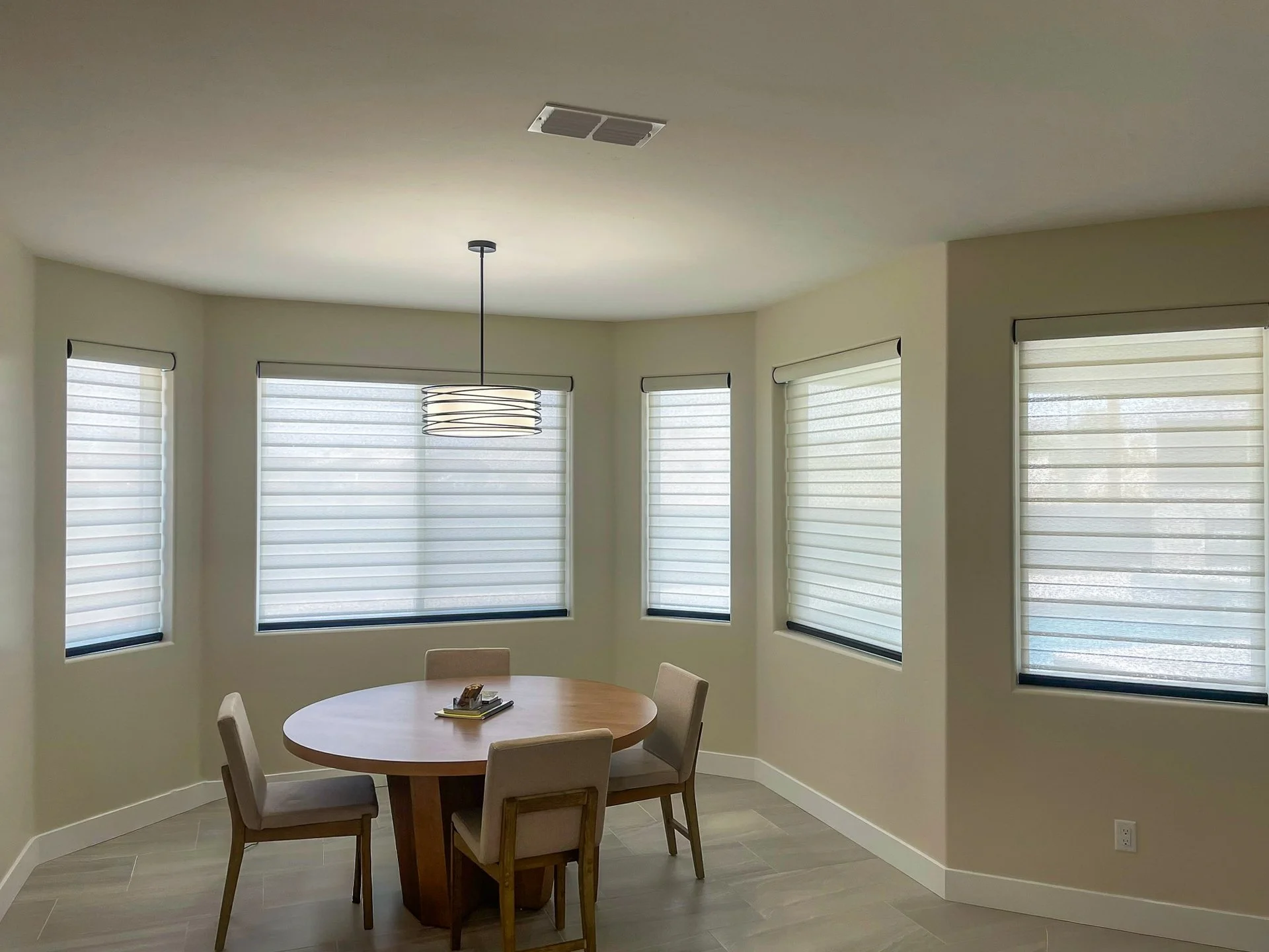 Round wooden dining table with four beige upholstered chairs in a bright room with five windows covered by white blinds and a modern hanging light fixture.