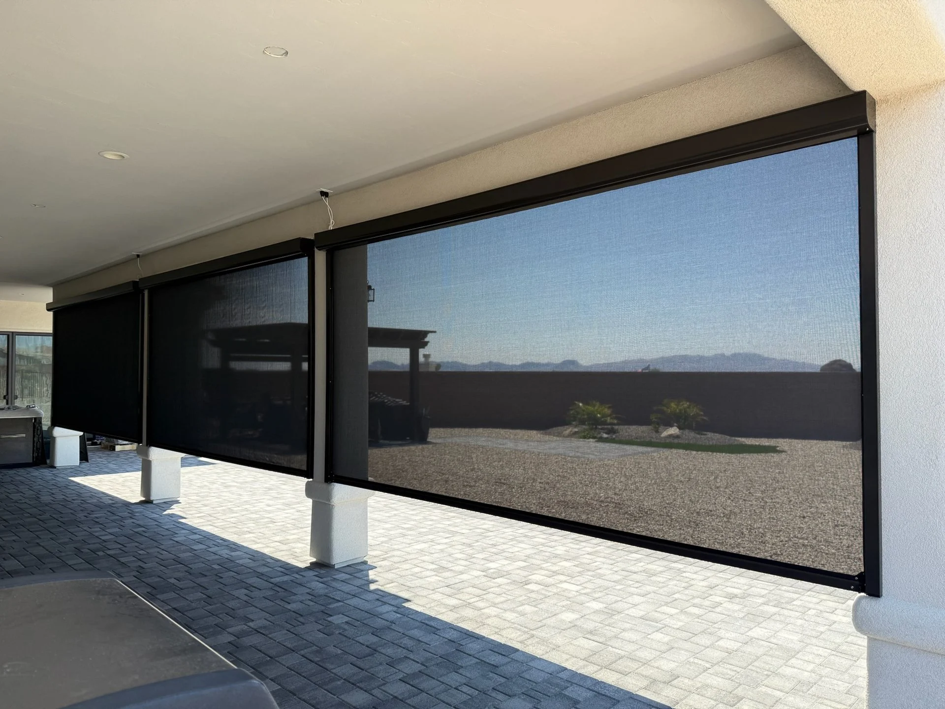 Outdoor patio with black retractable screens, view of gravel ground and distant mountains.