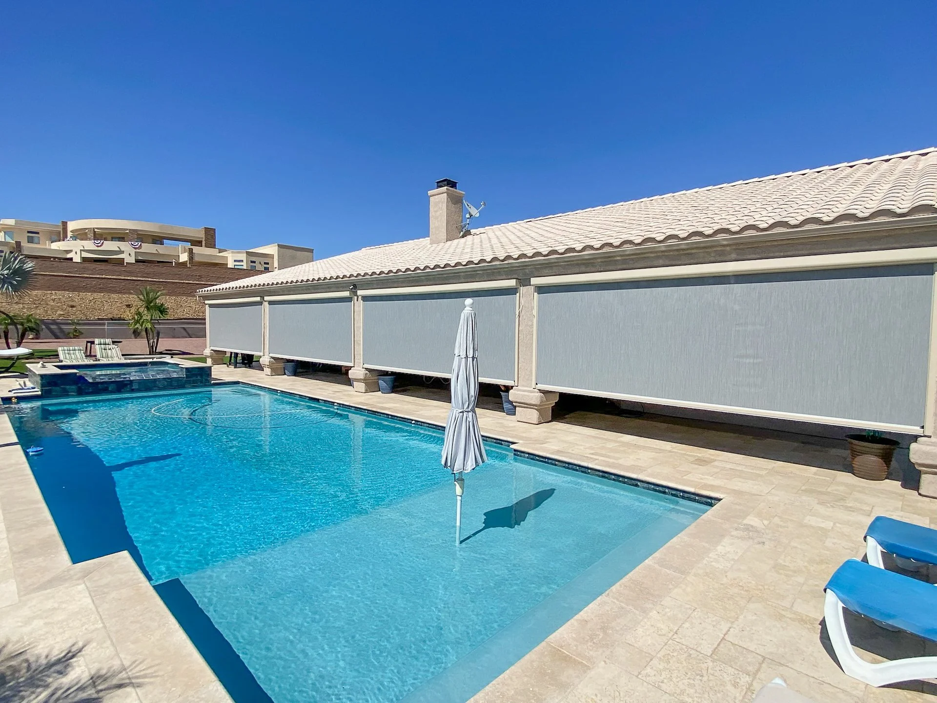 A backyard swimming pool with blue water, surrounded by beige tiles, with a white and blue umbrella and lounge chairs. There are some potted plants and a shaded structure with a covered privacy screen.