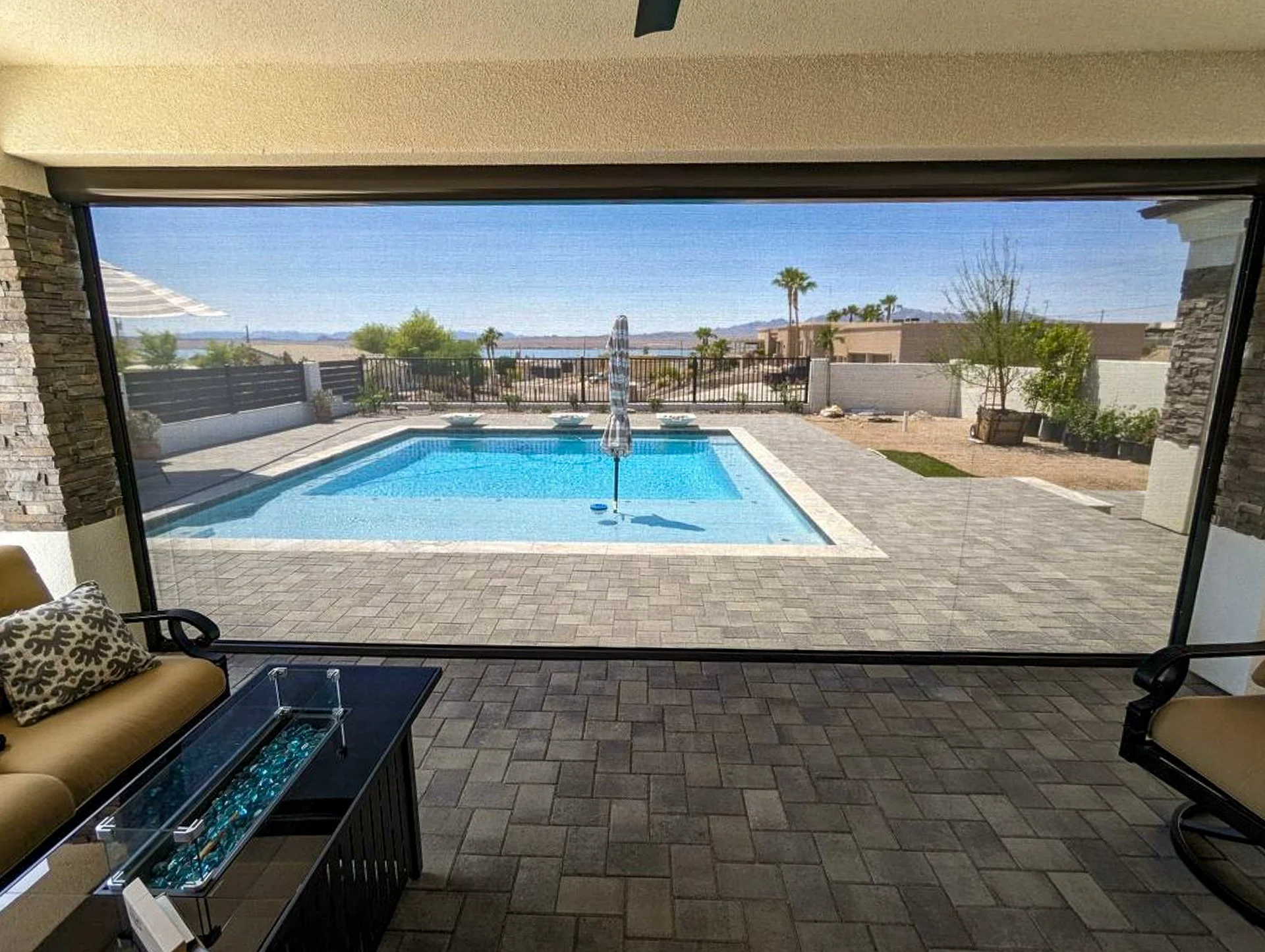 View of a backyard with a swimming pool, patio, and desert landscape through a large window.