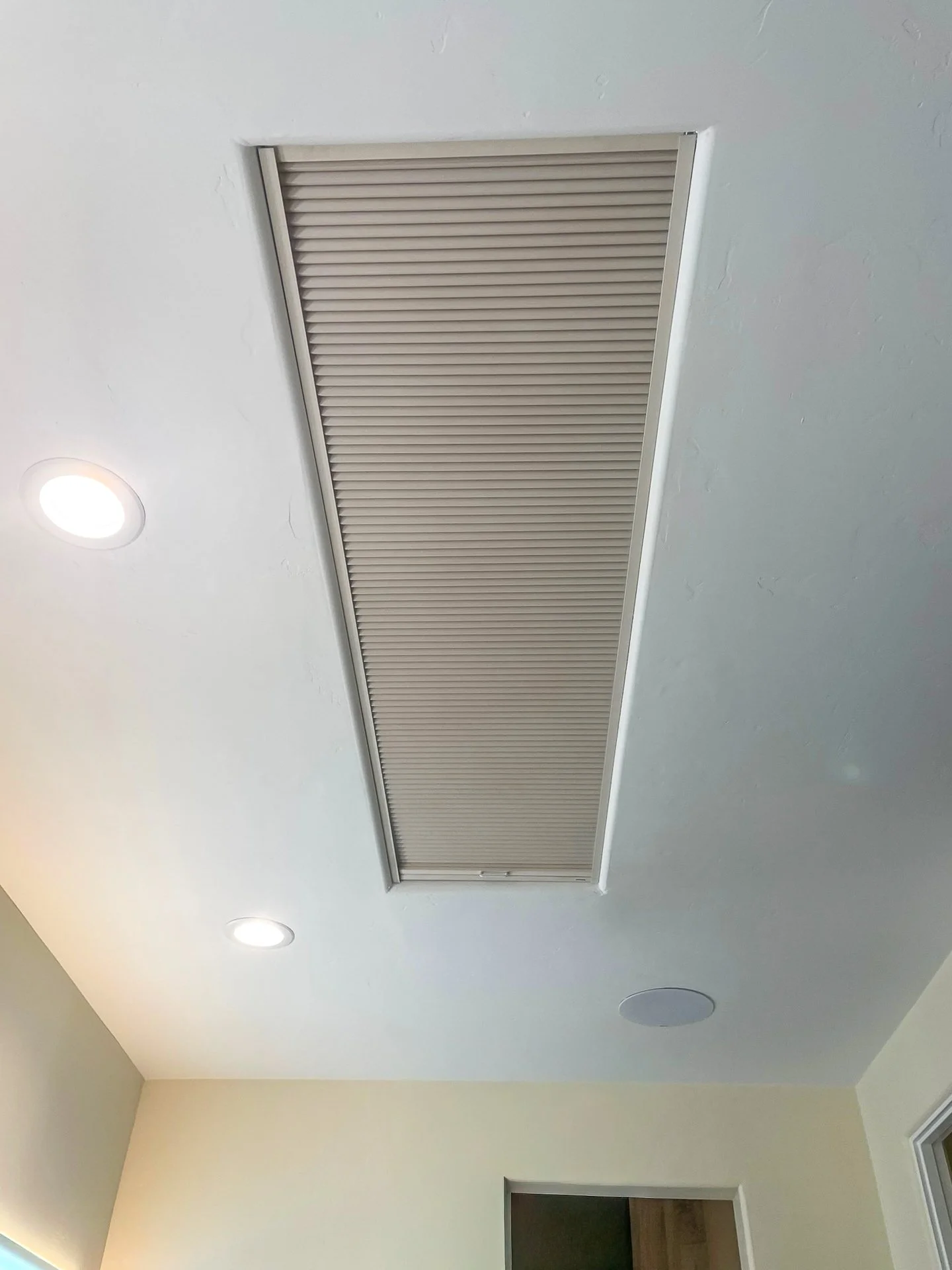 Ceiling vent or air duct with beige slats, surrounded by white ceiling with recessed lighting and a speaker, adjacent to yellow walls and a doorway.