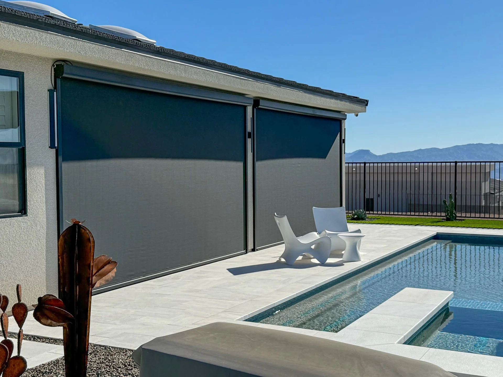A modern backyard with a swimming pool, white chairs, and a house with a sliding privacy screen. Mountains are visible in the distance under a clear blue sky.