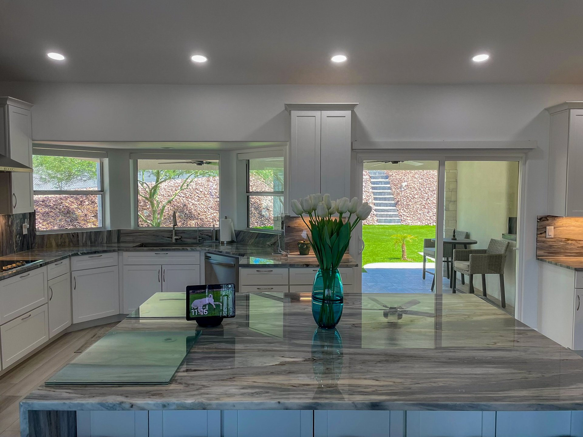 Interior view of a modern kitchen with white cabinetry, a marble countertop, large windows, and a sliding glass door leading to a backyard with green grass, steps, and outdoor seating. A blue vase with white tulips is on the countertop, and a tablet 