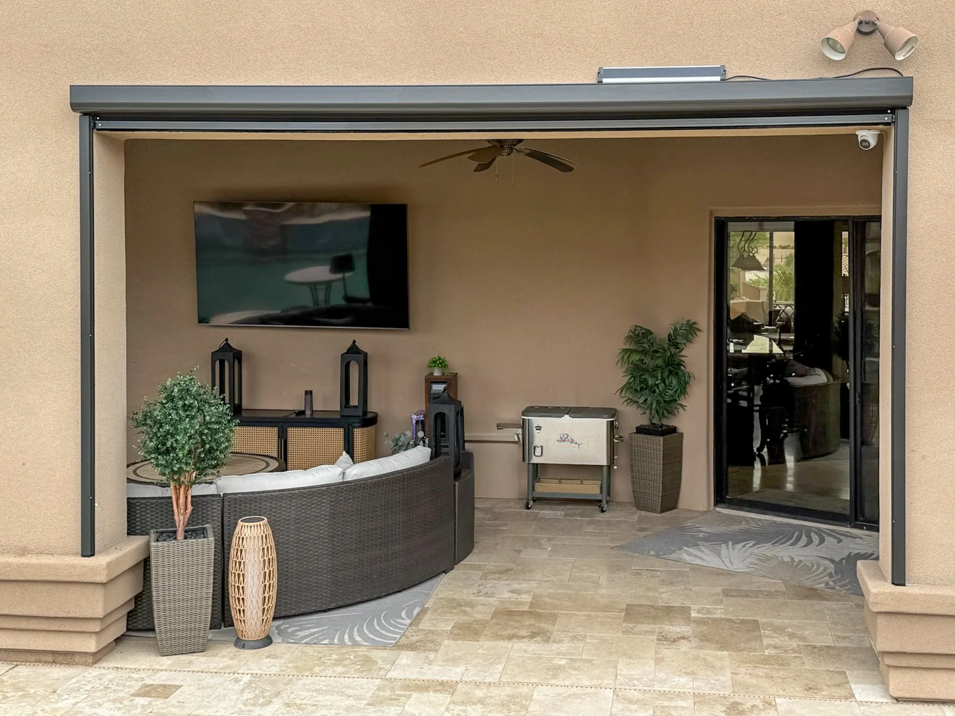 Outdoor patio with a wall-mounted TV, potted plants, wicker furniture, a small grill, and a sliding glass door leading inside a house.