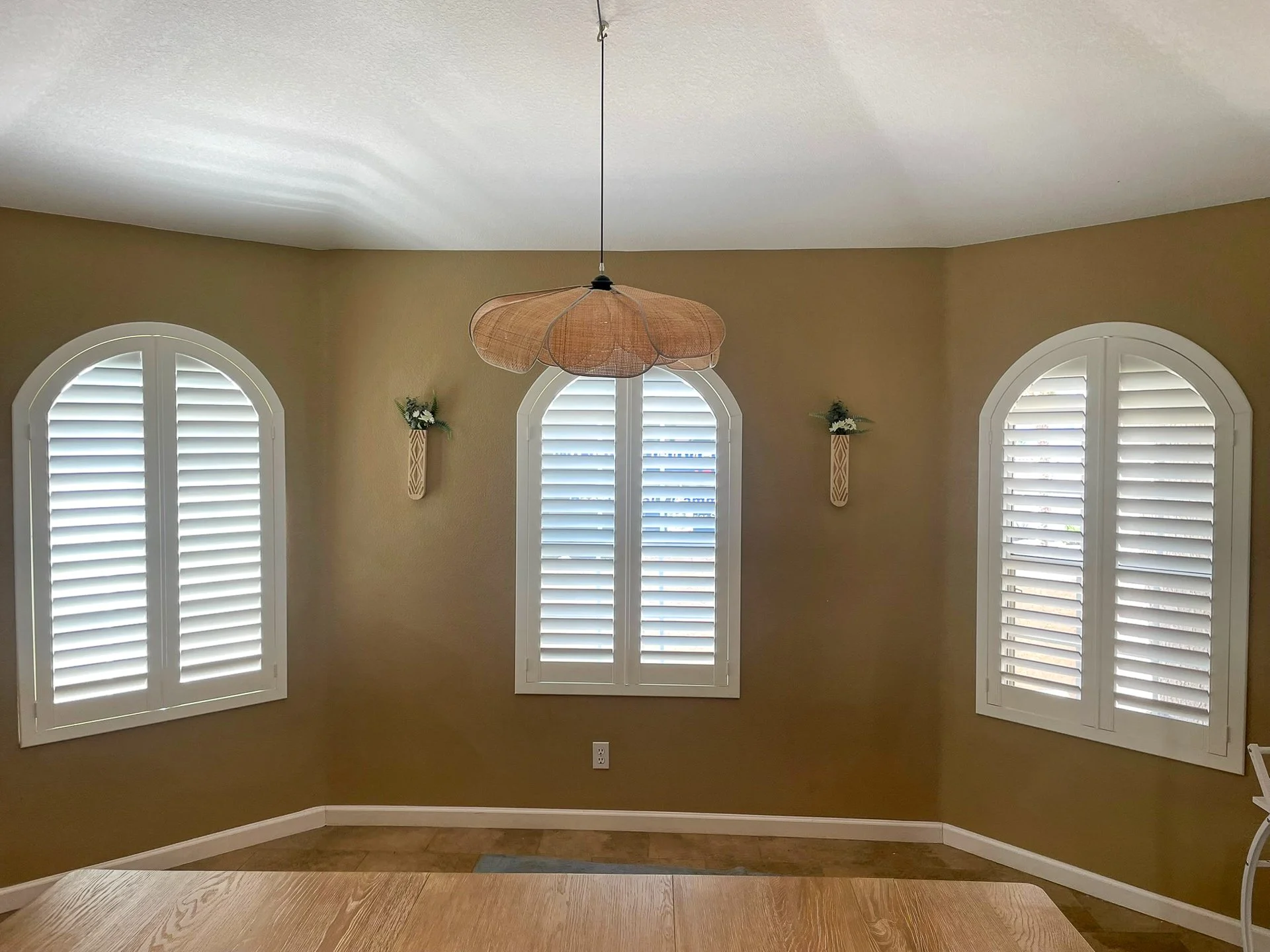 A room with three arched windows with white shutters on a beige wall, a wooden table in the foreground, and a brown pendant light hanging from the ceiling.