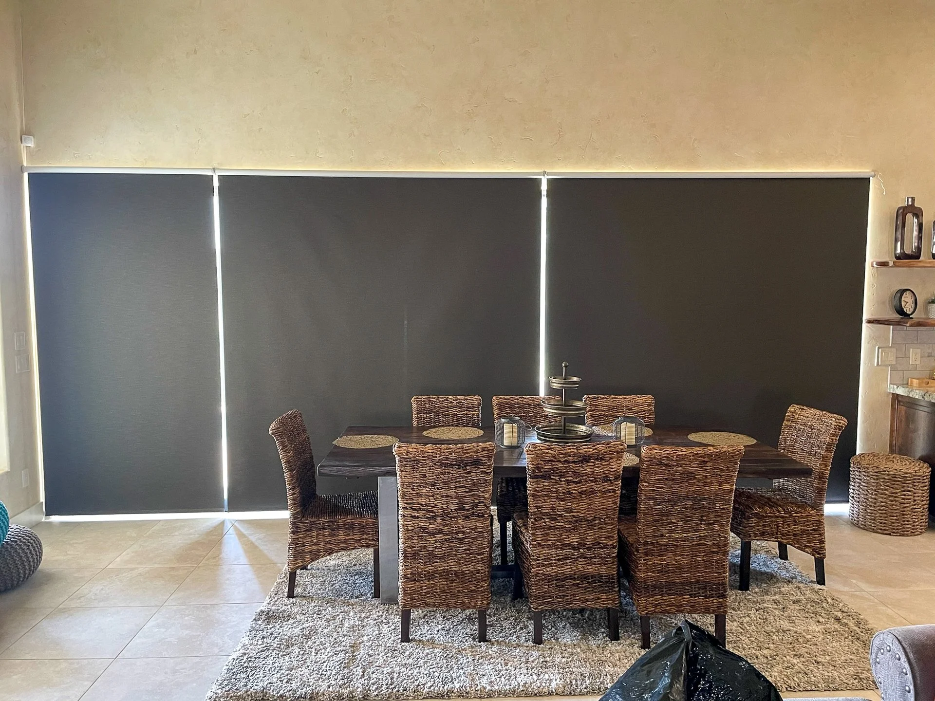 A dining room with a dark wooden table, eight wicker chairs, a sunken chandelier, and black window blinds covering a large window, with a beige textured wall.