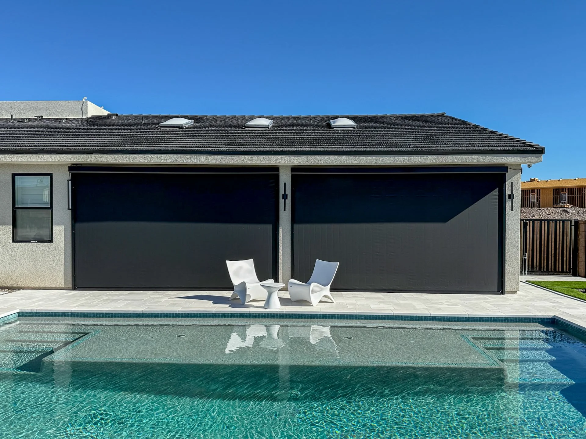 Modern house with a swimming pool, two white chairs, and a small table on the patio under a clear blue sky.